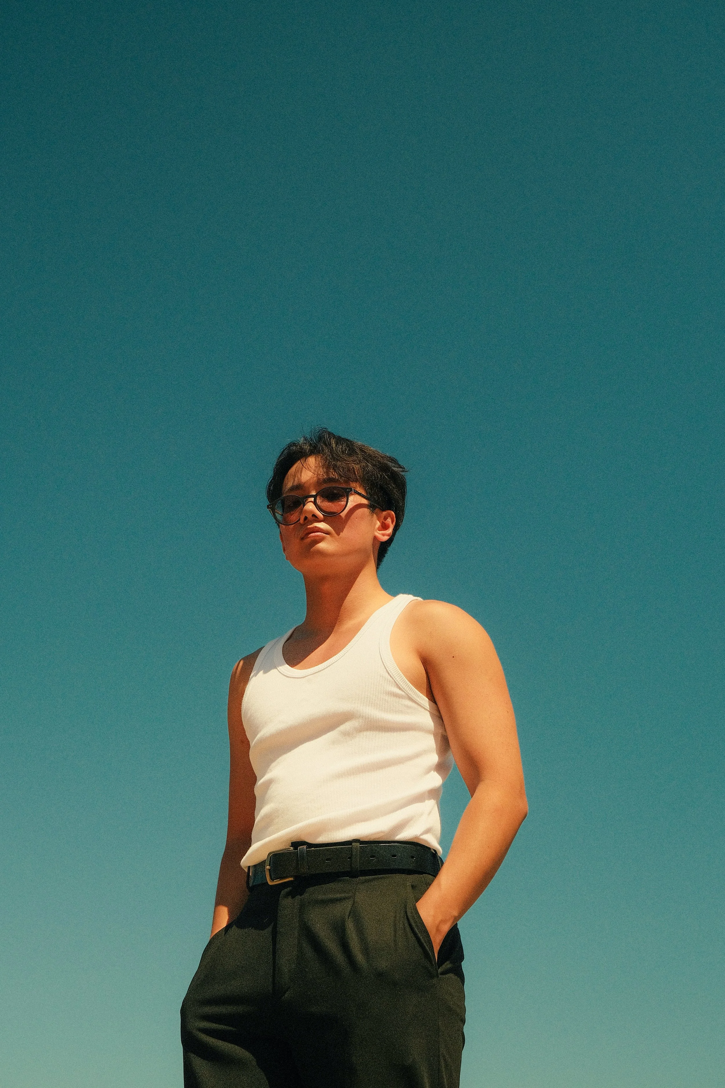 Une personne portant un débardeur blanc, un pantalon noir et des lunettes, se tenant sous un ciel bleu clair.
