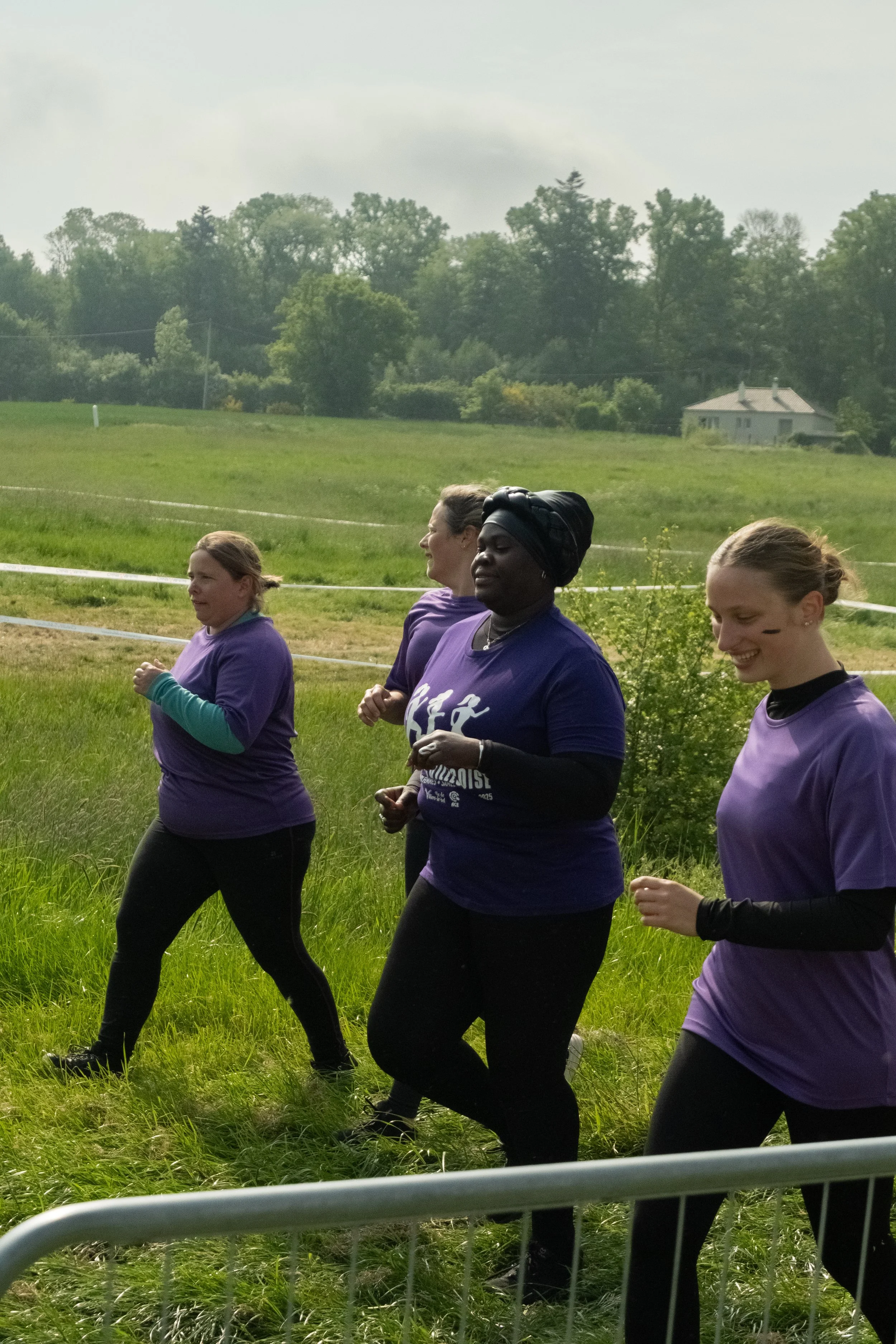 Groupe de quatre femmes qui courent ensemble dans un champ vert, portant des t-shirts violets, dans un environnement naturel avec des arbres et une maison en arrière-plan.