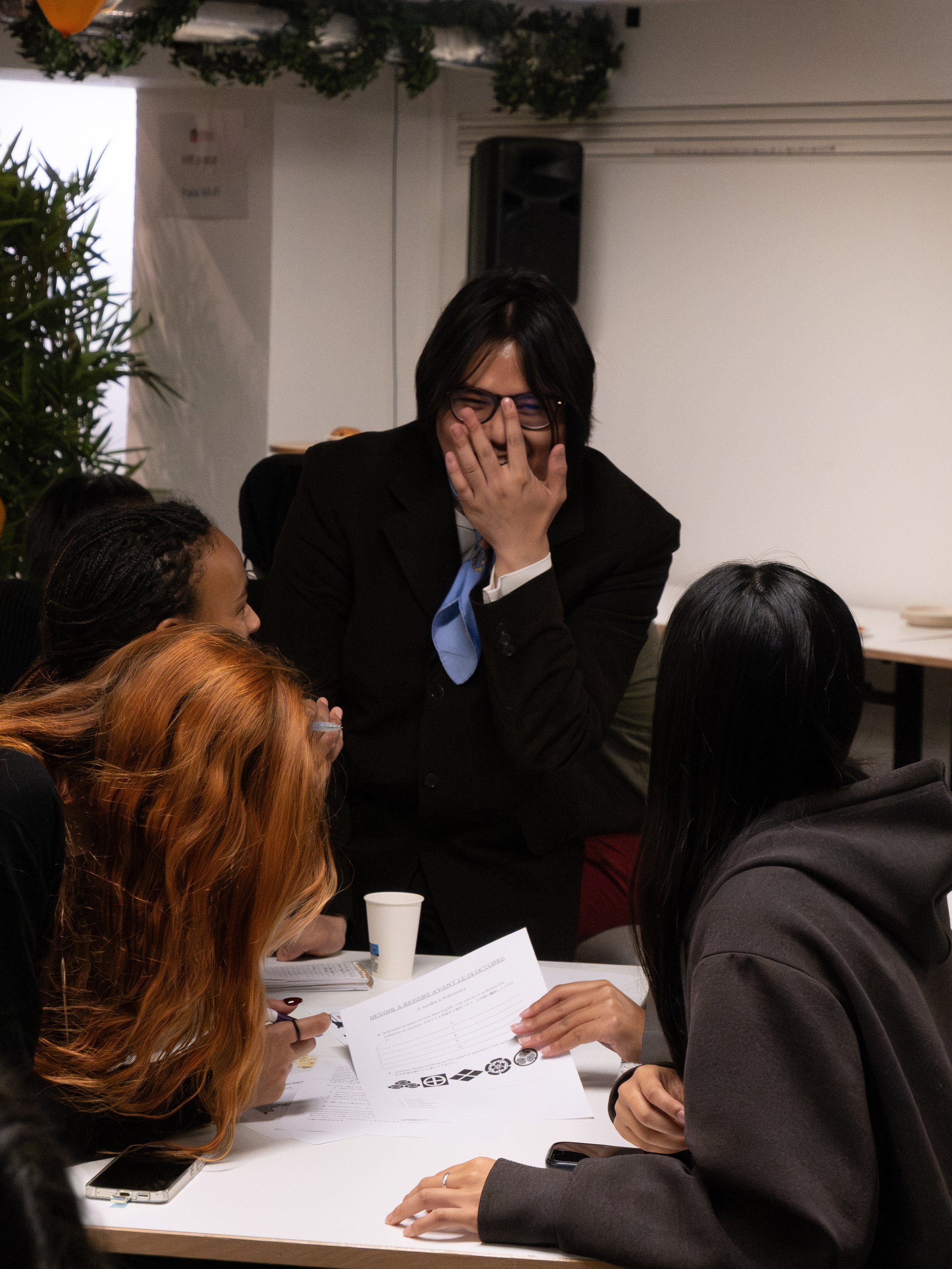 Groupe de jeunes femmes autour d'une table, en pleine conversation, avec une femme en costume qui rit et couvre sa bouche, dans un environnement intérieur.