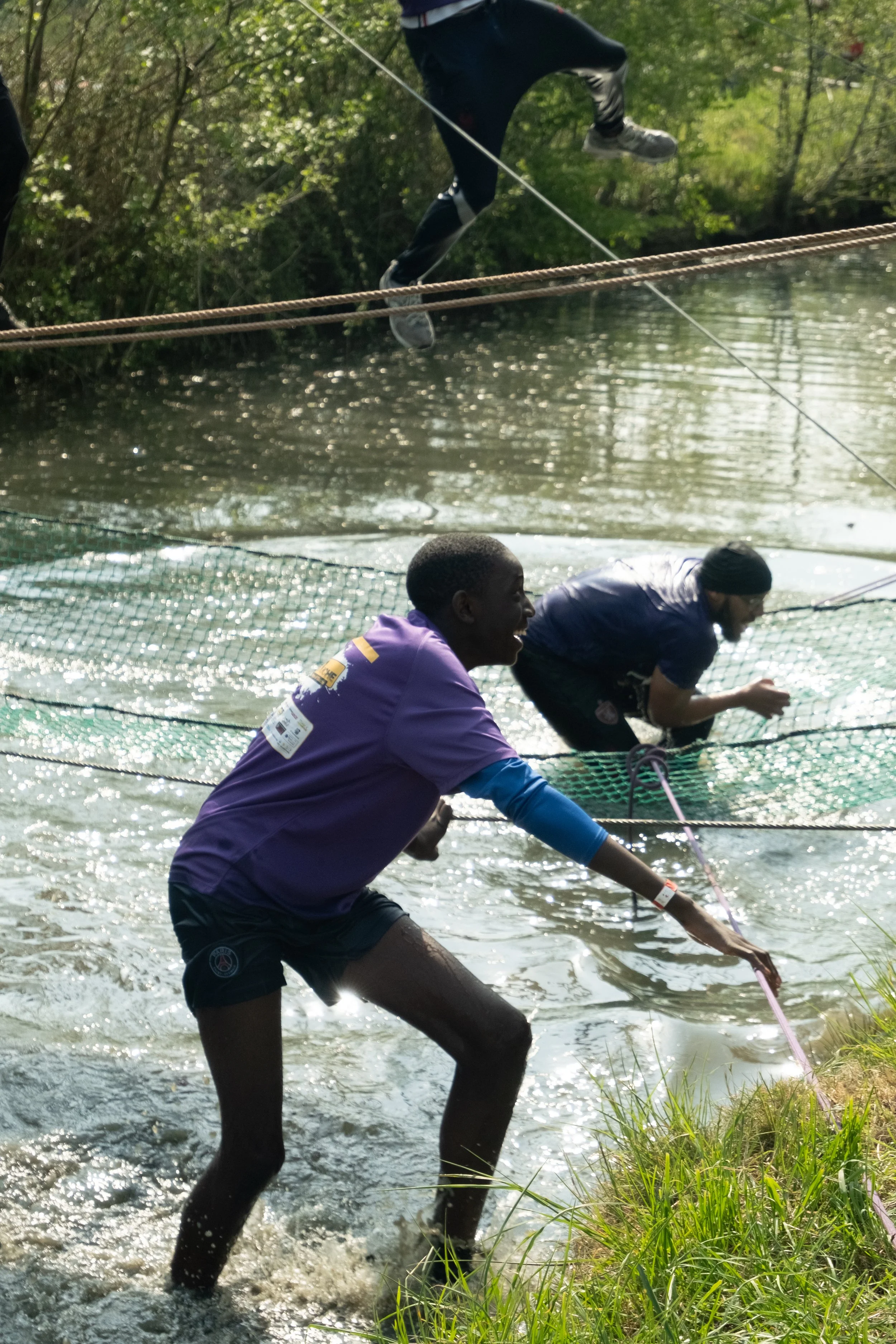 Deux jeunes hommes jouent à la pêche dans une rivière, l'un d'eux est vêtu d'un maillot violet et remue dans l'eau, l'autre en arrière est plié au sol, tous deux pêchent à la ligne sous un pont en bois au fond d'une forêt dense.