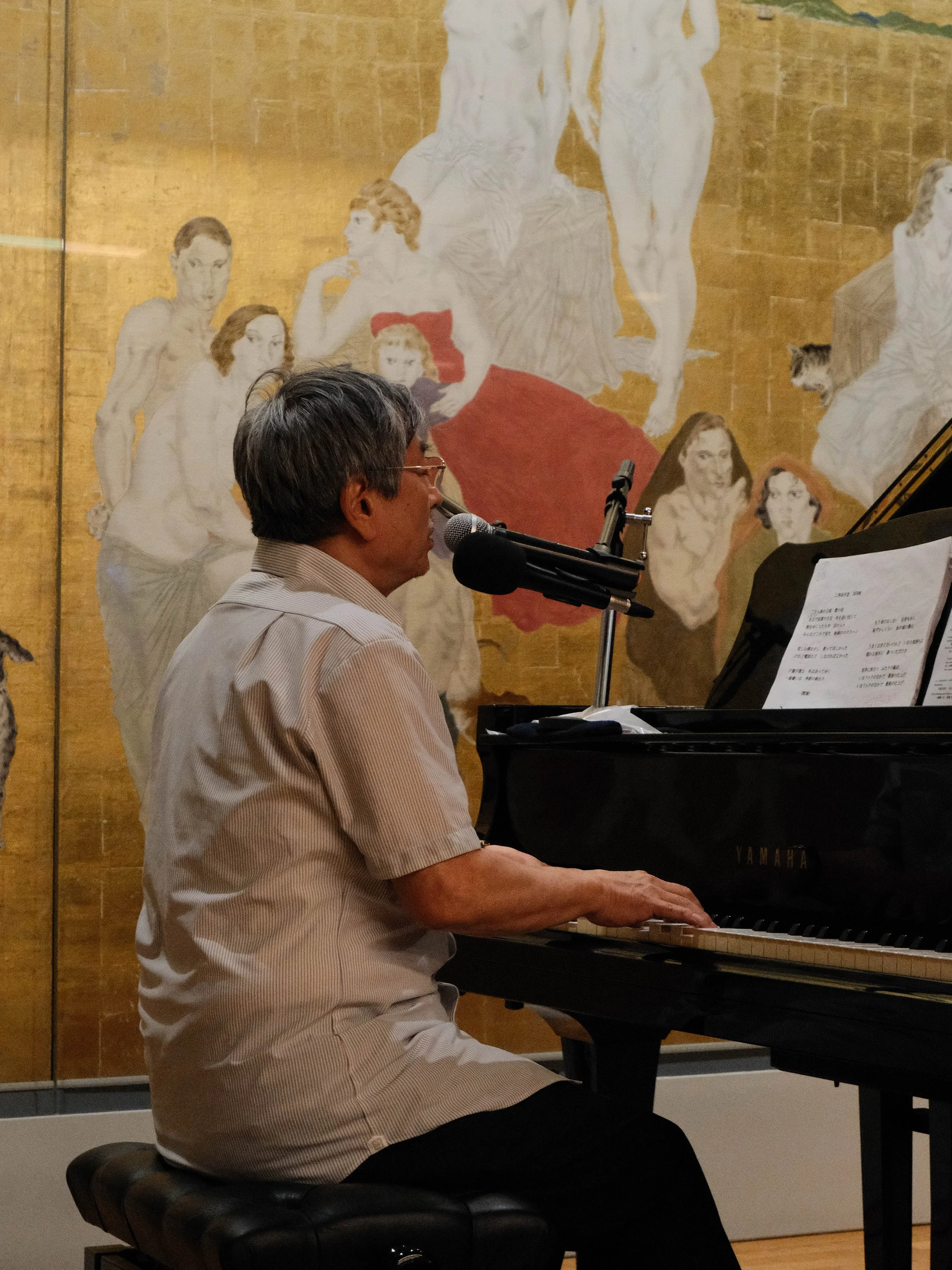 Un homme jouant du piano à queue noir, chantant ou parlant dans un microphone, avec un tableau mural derrière lui représentant plusieurs femmes nues et une femme en rouge couché, dans une ambiance intérieure.