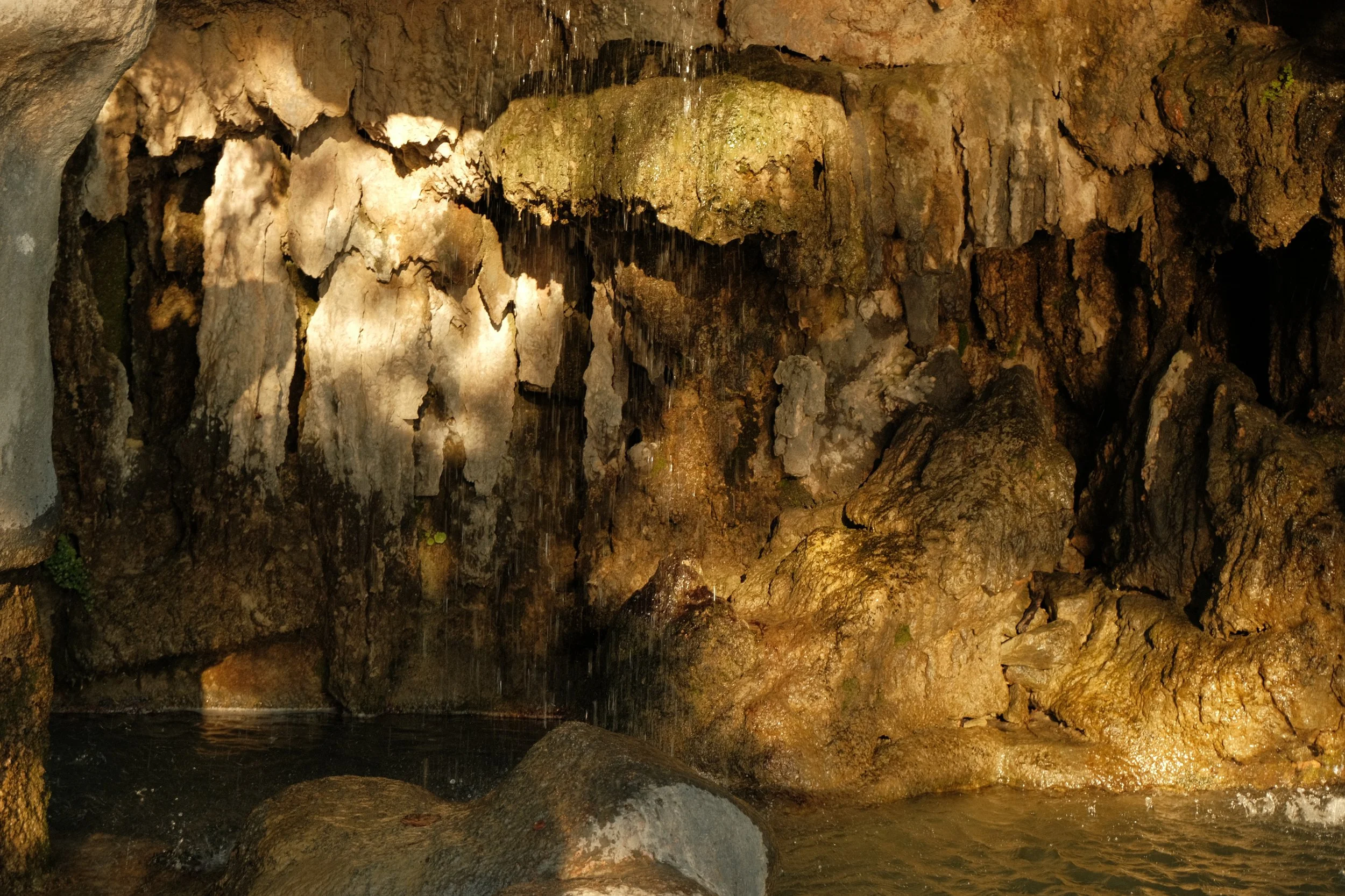 Intérieur d'une grotte avec des rochers, une petite cascade d'eau tombant sur les rochers, et une étendue d'eau au sol.
