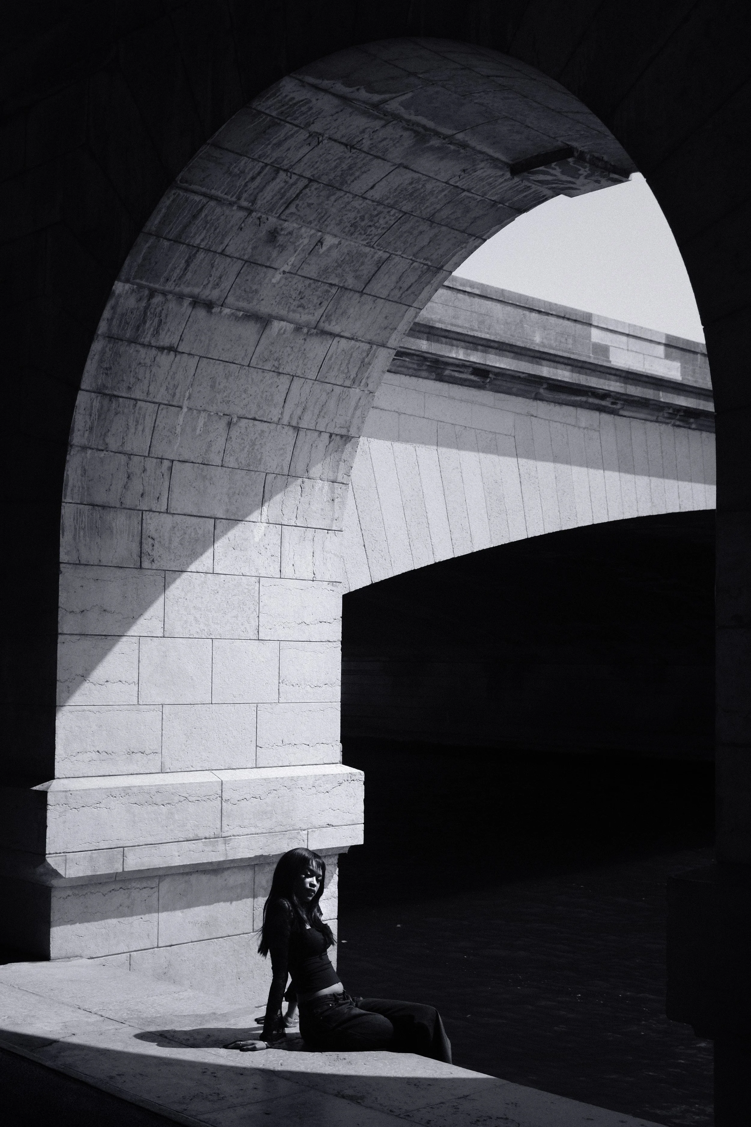 Une femme assise sous un pont en pierre, en noir et blanc, avec une lumière qui crée des ombres et des contrastes visible sur le mur et au sol.