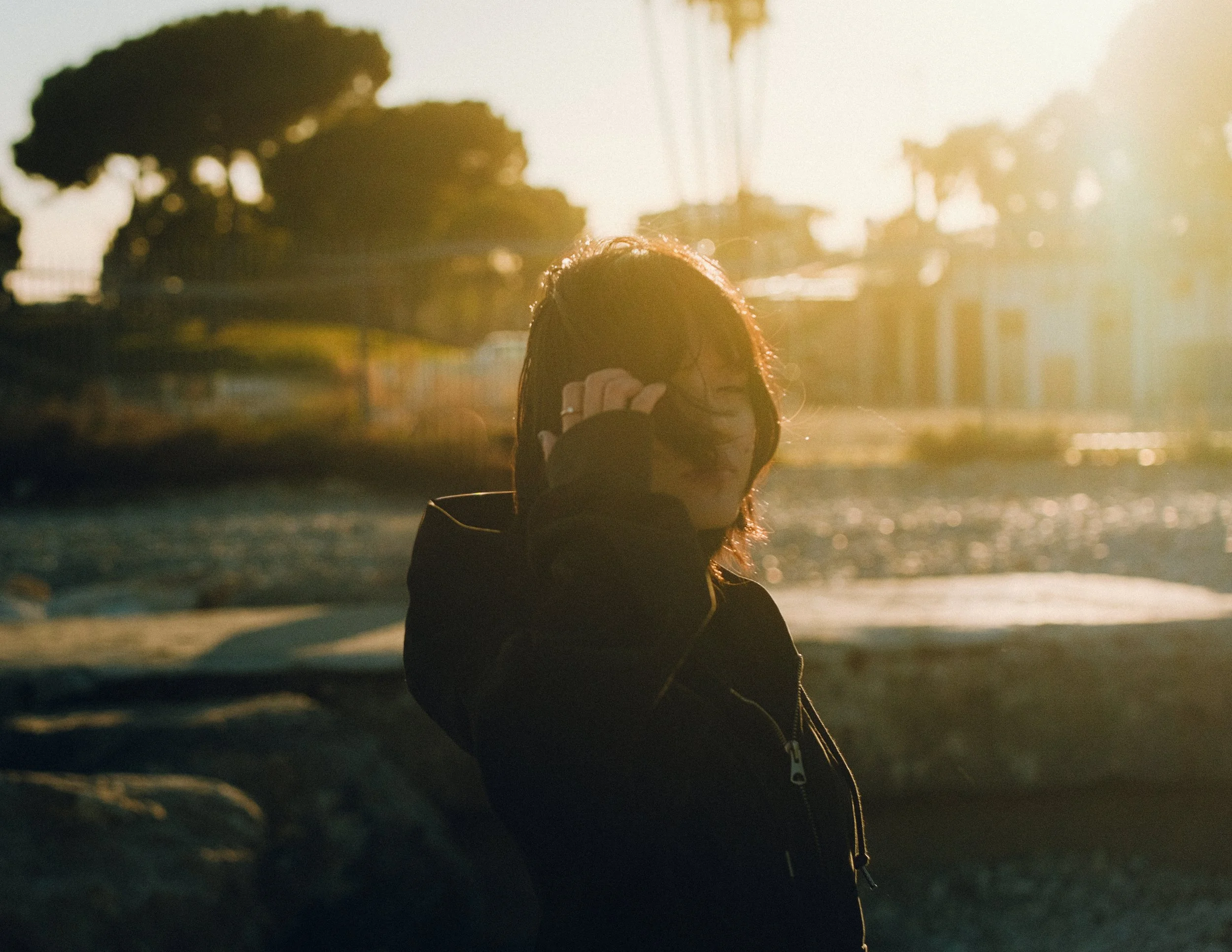 Une personne avec des cheveux foncés et une veste noire, se tenant près d'une rivière ou d'un plan d'eau au coucher du soleil, portant des lunettes de soleil sur la tête.