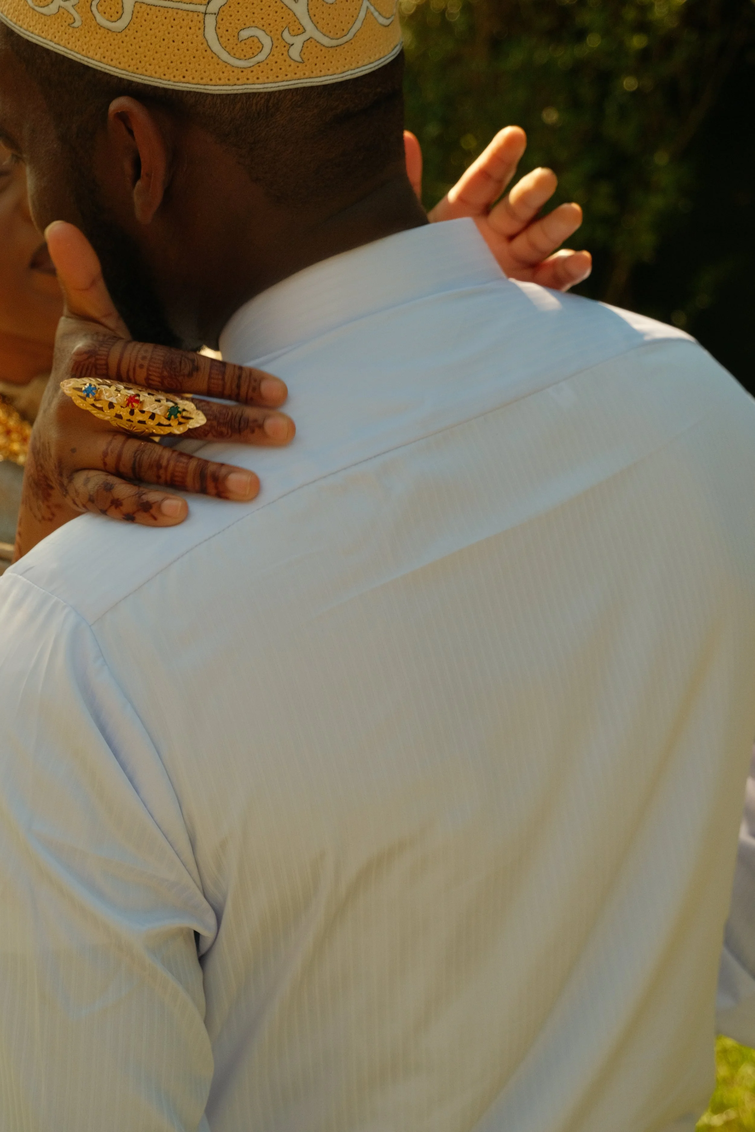 Un homme portant une chemise blanche, vu de dos, avec une femme dont la main repose sur son épaule. La femme porte une bague dorée décorée de pierres colorées, et a des tatouages sur les doigts. L'arrière-plan est un espace extérieur avec des arbres.