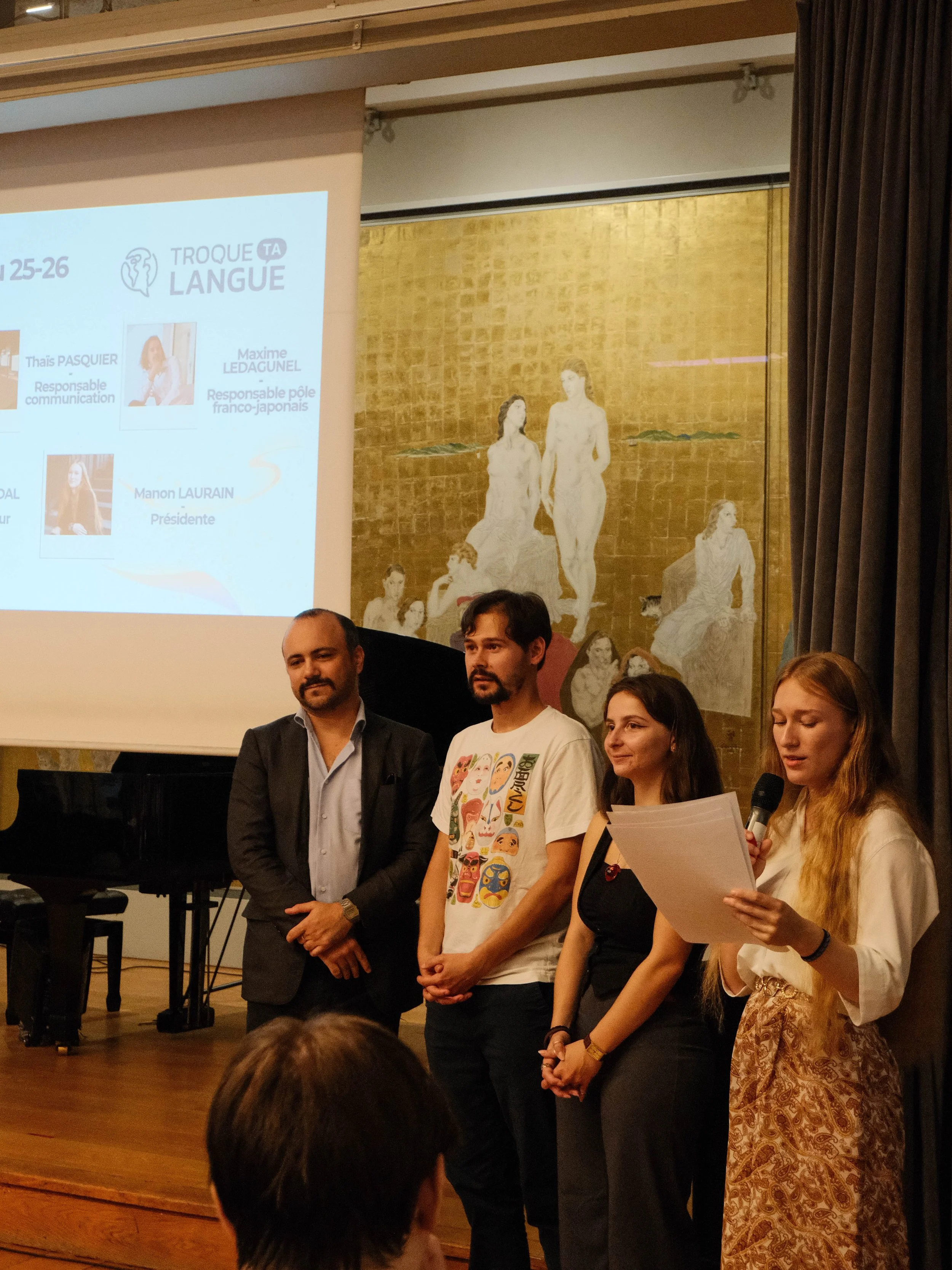 Quatre personnes debout devant un écran projetant une présentation lors d'une conférence ou d'un événement. La femme à droite lit un discours en tenant un microphone et des papiers. Le fond montre une peinture dorée avec des figures humaines.