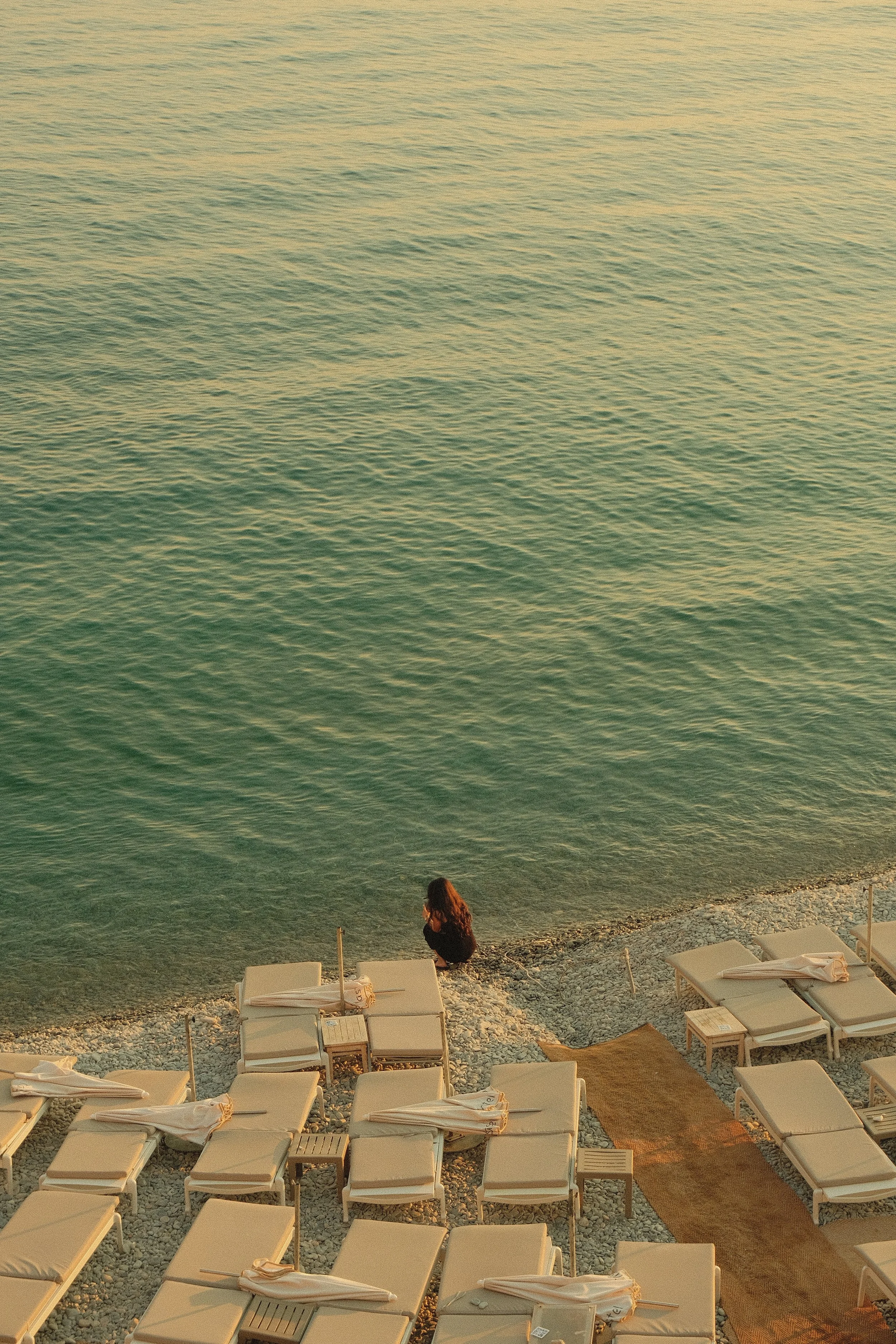 Une personne assise sur la plage au bord de la mer avec des chaises longues et des parasols pliés.