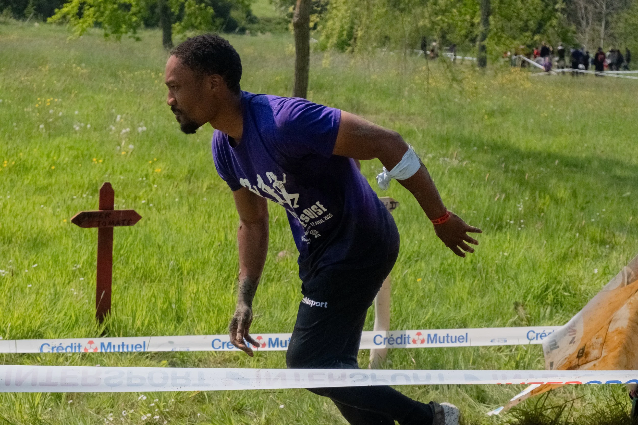 Un homme participe à une course en plein air, il est courbé en avant, portant un T-shirt violet et un pantalon noir, avec une bande blanche autour du bras, dans un champ vert avec des arbres en arrière-plan.