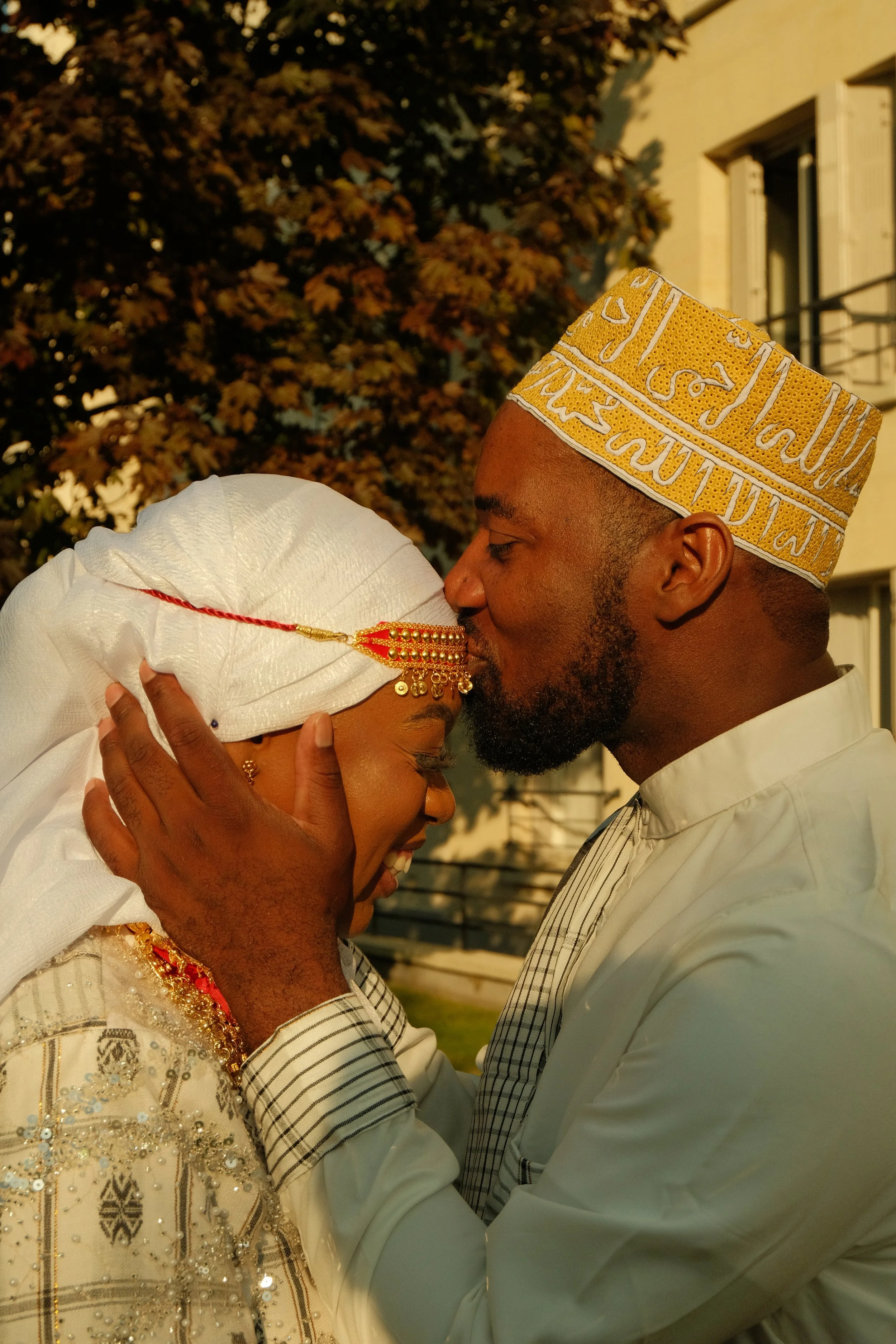 Un couple en habit traditionnel en train de s'embrasser tendrement lors d'une cérémonie nuptiale, avec une femme portant un voile blanc décoré de bijoux dorés et un homme portant un chapeau traditionnel brodé.