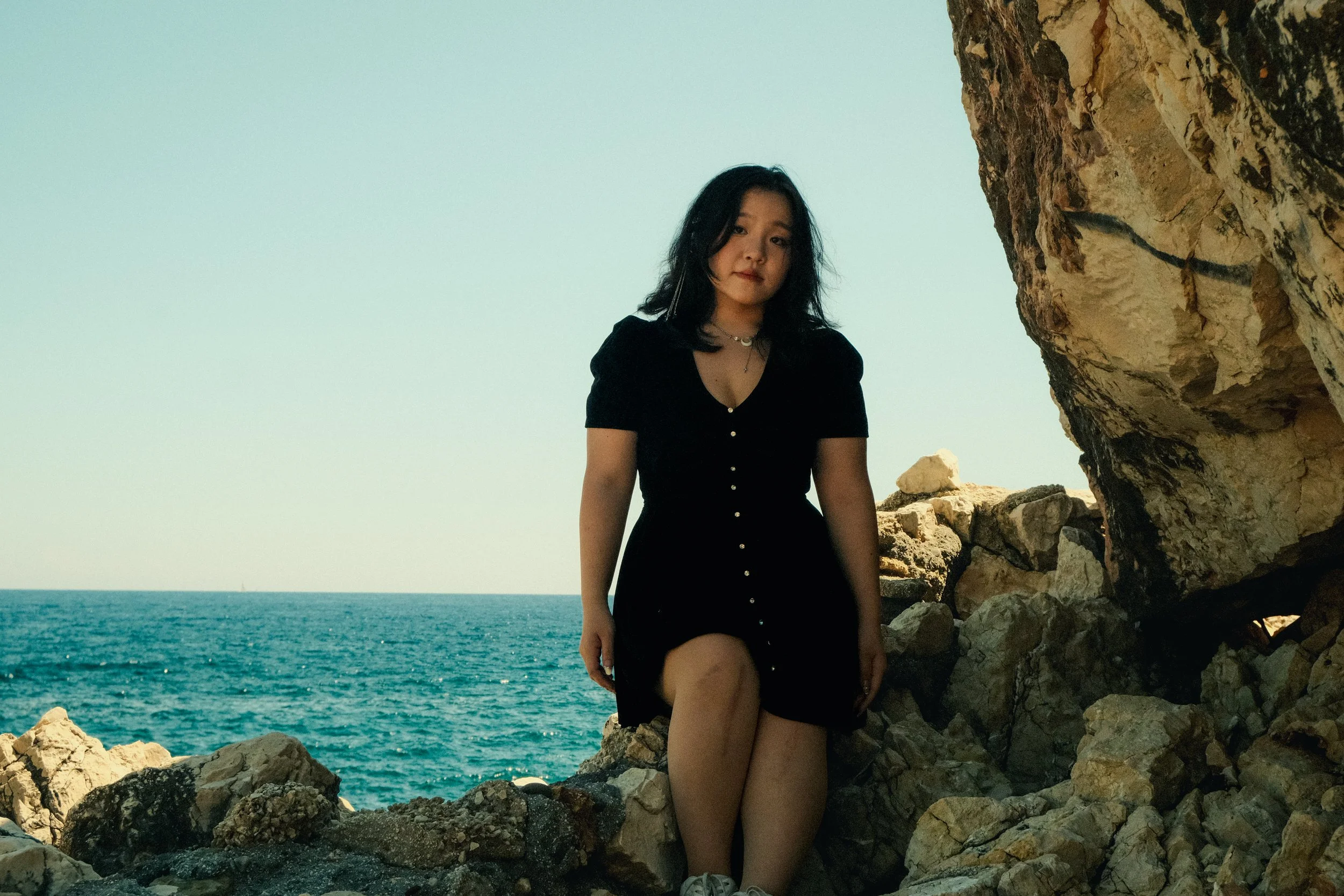 Jeune femme assise entre des rochers près de la mer, portant une robe noire, en regardant vers l'objectif, avec la mer et un ciel clair en arrière-plan.
