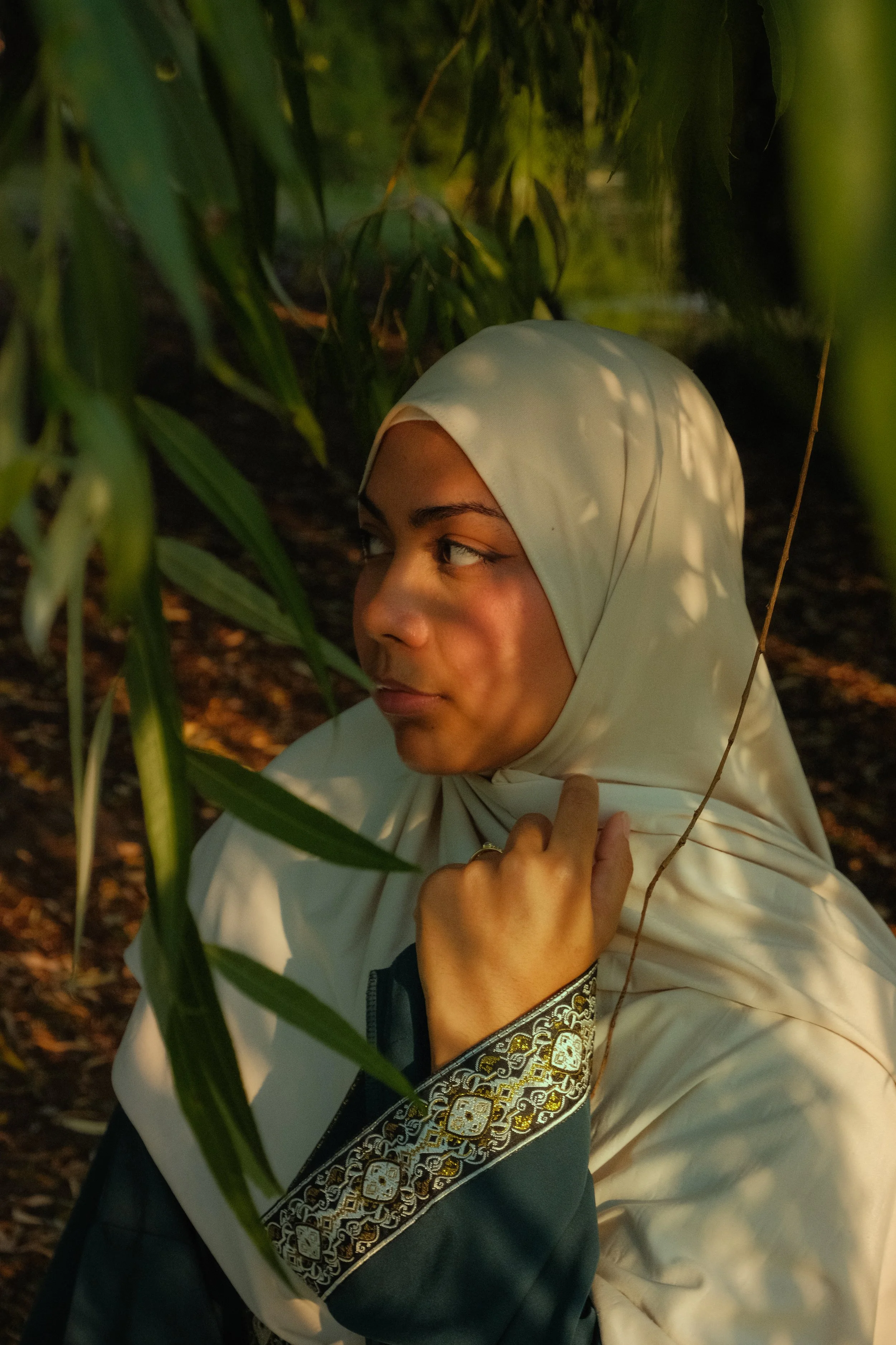 Femme en hijab blanc parmi des feuilles vertes, dans un environnement naturel, avec des jeux d'ombres et de lumières sur son visage.