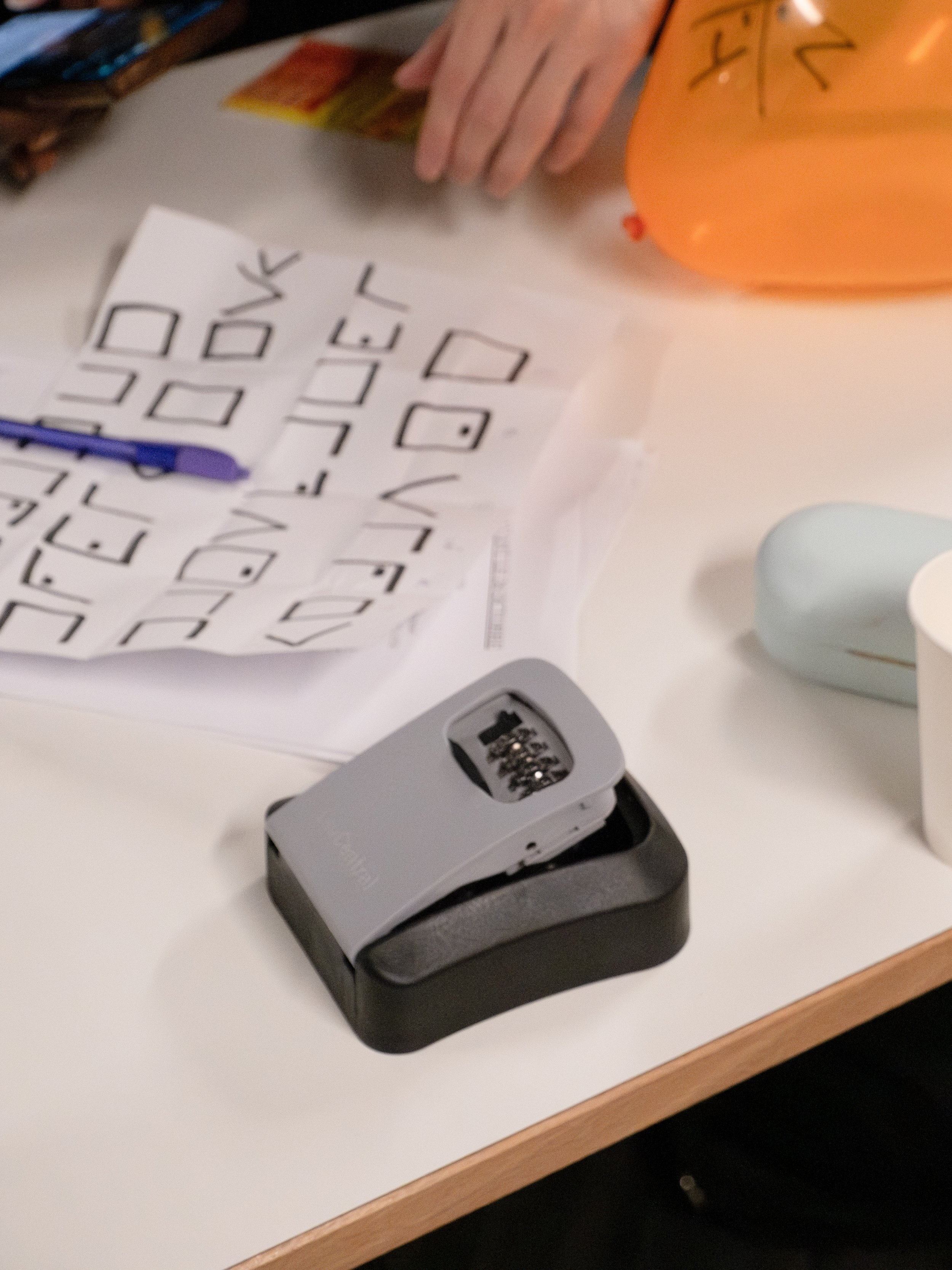 Une planche avec des dessins géométriques et un stylo bleu, une main tenant une feuille, une machine à relier, un ballon orange, un album d'images en arrière-plan, et un bloc de papier blanc sur une table.