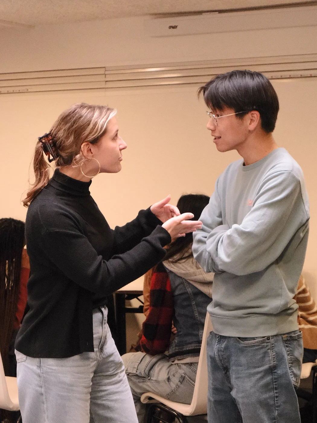 Une femme et un homme discutent face à face dans une pièce, d'autres personnes sont assises en arrière-plan.