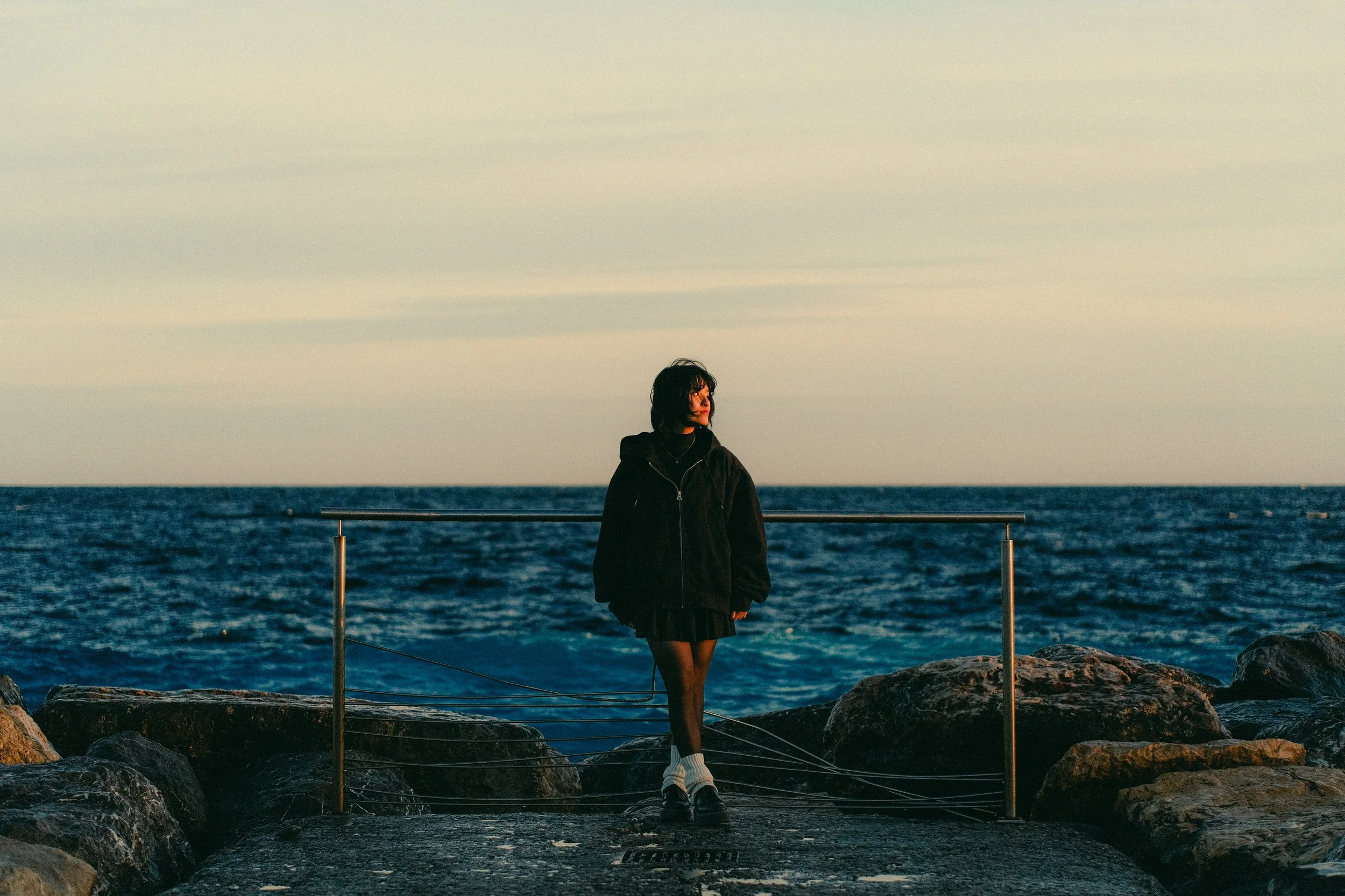 Une personne debout sur un quai en pierre avec des rochers, regardant vers la mer au coucher du soleil, portant un manteau noir et des chaussures blanches.
