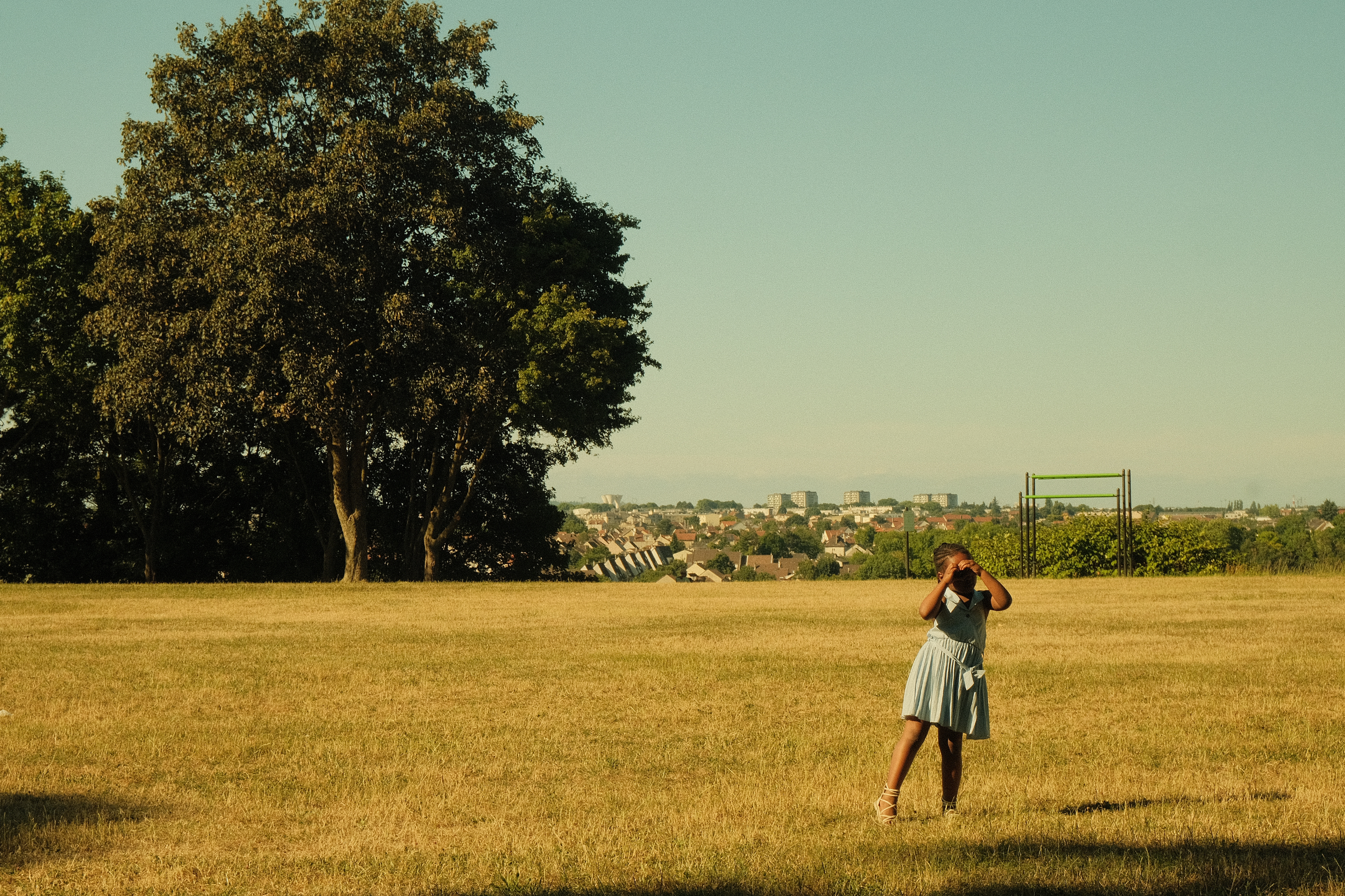 Une jeune fille en robe bleue se tient dans une grande étendue de pelouse, portant un masque et prenant une photo avec un appareil photo ou un téléphone. En arrière-plan, il y a de grands arbres et une vue sur une ville ou un lotissement, sous un cie