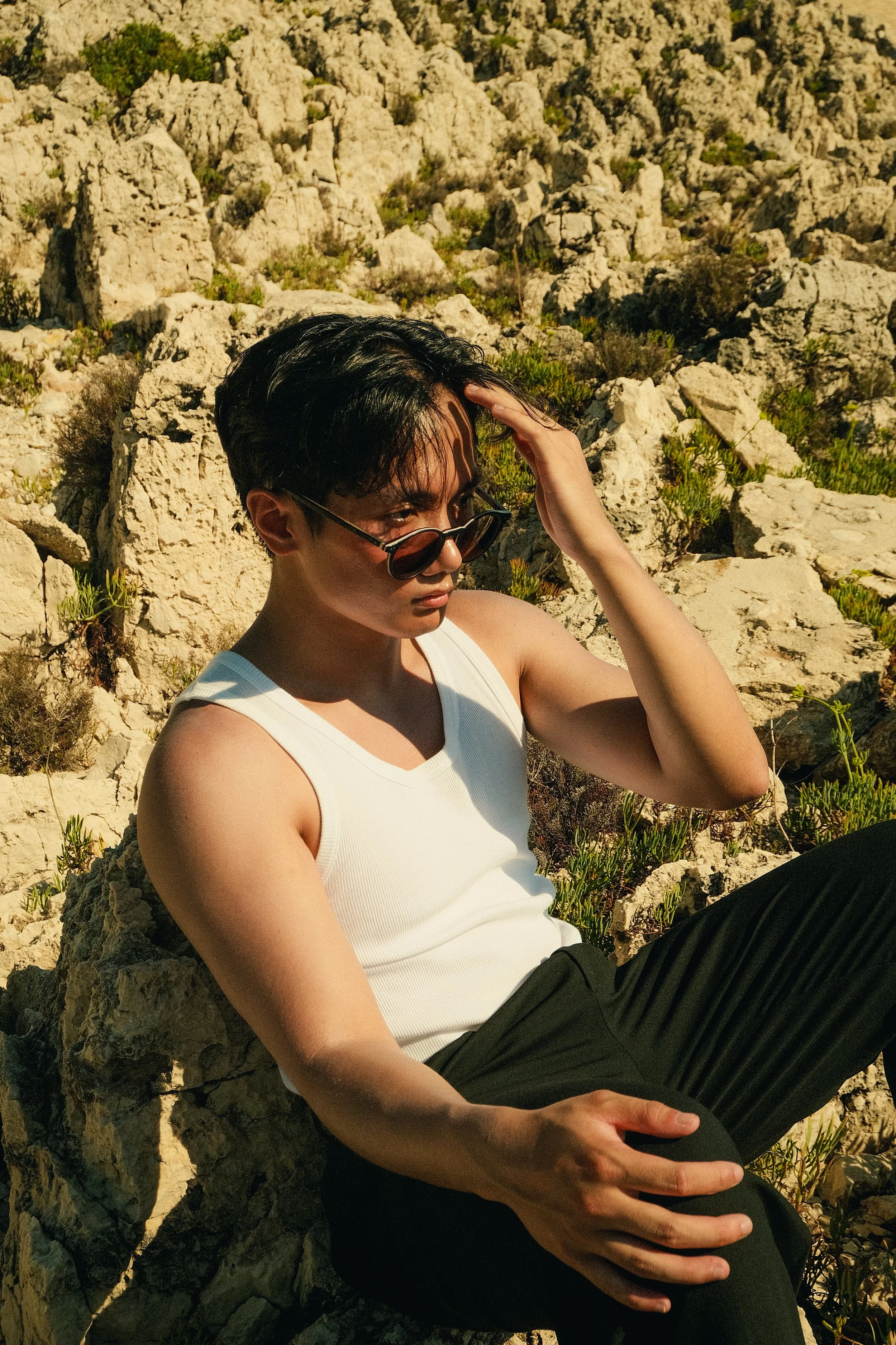 Jeune homme assis sur des rochers, portant un débardeur blanc et des lunettes de soleil, dans un paysage rocheux ensoleillé.