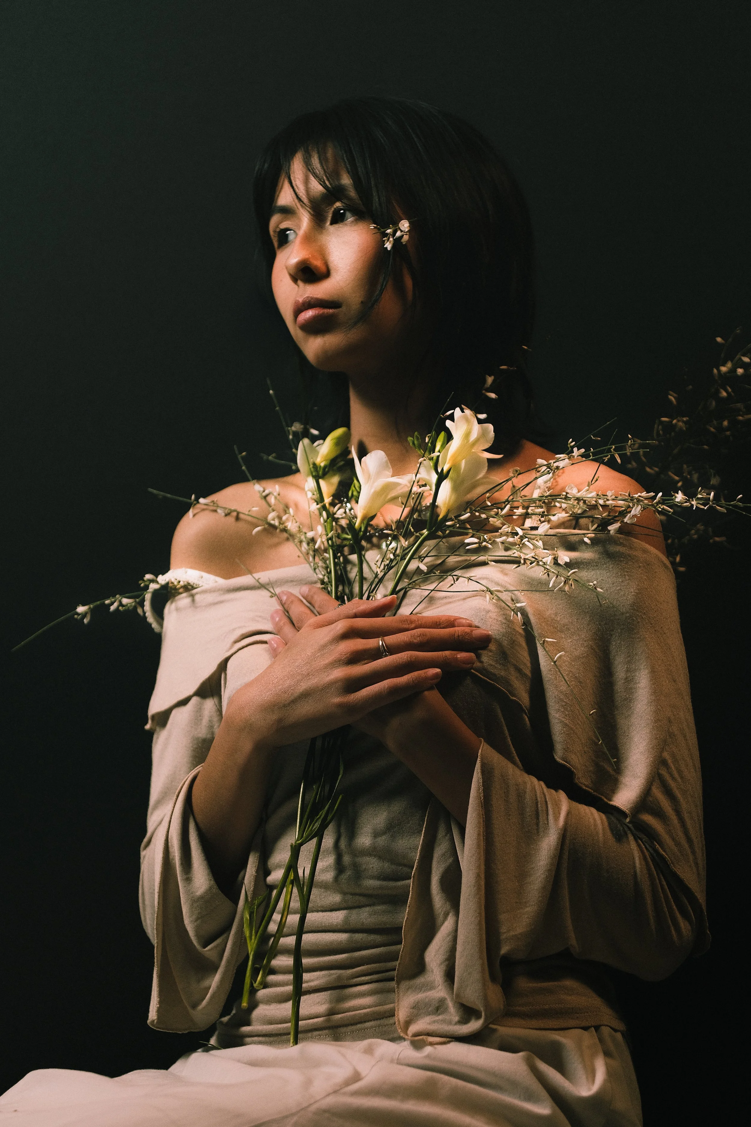 Une femme avec une robe beige tient un bouquet de fleurs blanches et de branches, regardant pensivement vers la gauche, contre un fond sombre.