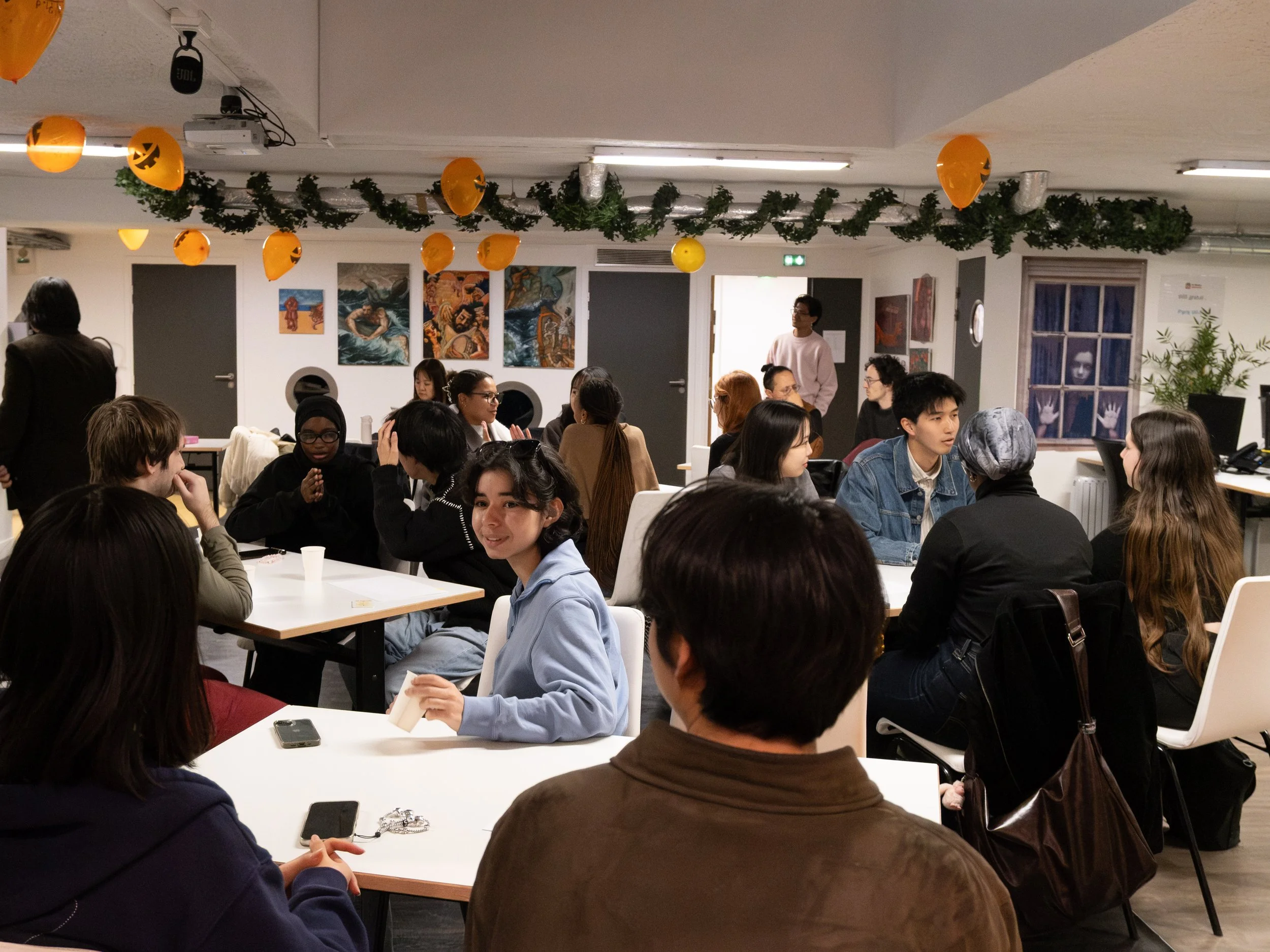 Groupe de personnes discutant dans une salle décorée de ballons orange Halloween et guirlandes vertes, avec des œuvres d'art sur les murs.