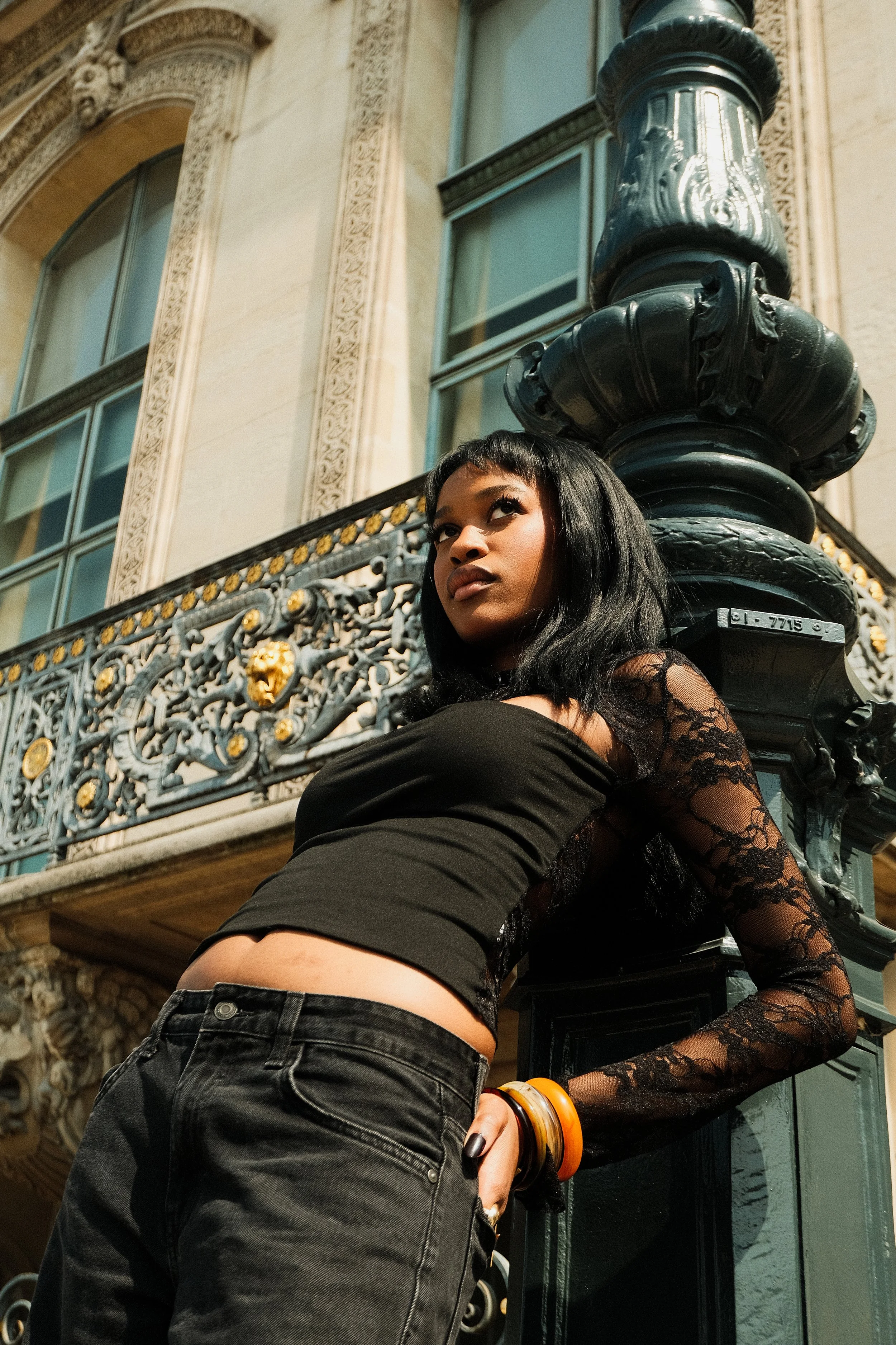 Jeune femme noire posant près d'un lampadaire en fer forgé à l'extérieur d'un bâtiment architectural en pierre, portant un haut noir en dentelle et un jean noir.
