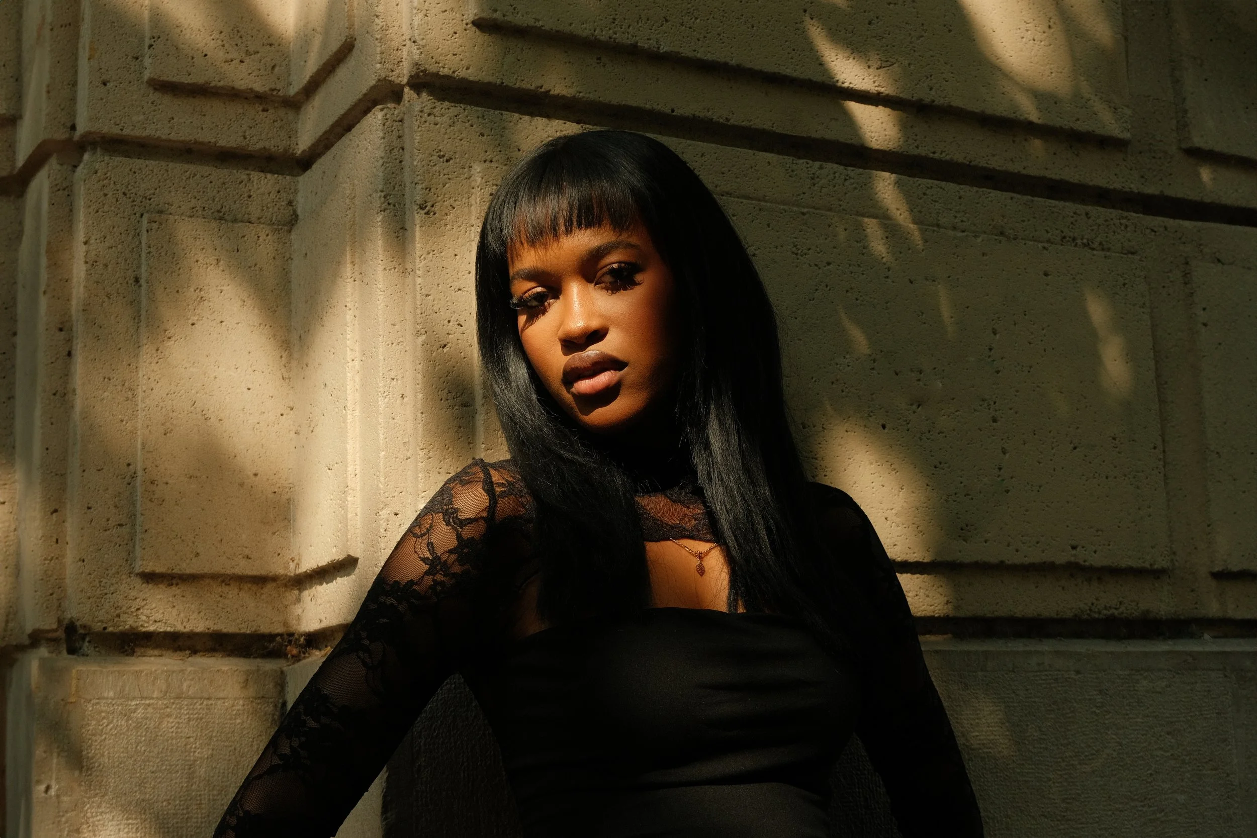 Femme avec long noir et décolletr en dentelle noire, assise contre un mur en pierre, avec des ombres de feuilles sur le mur.