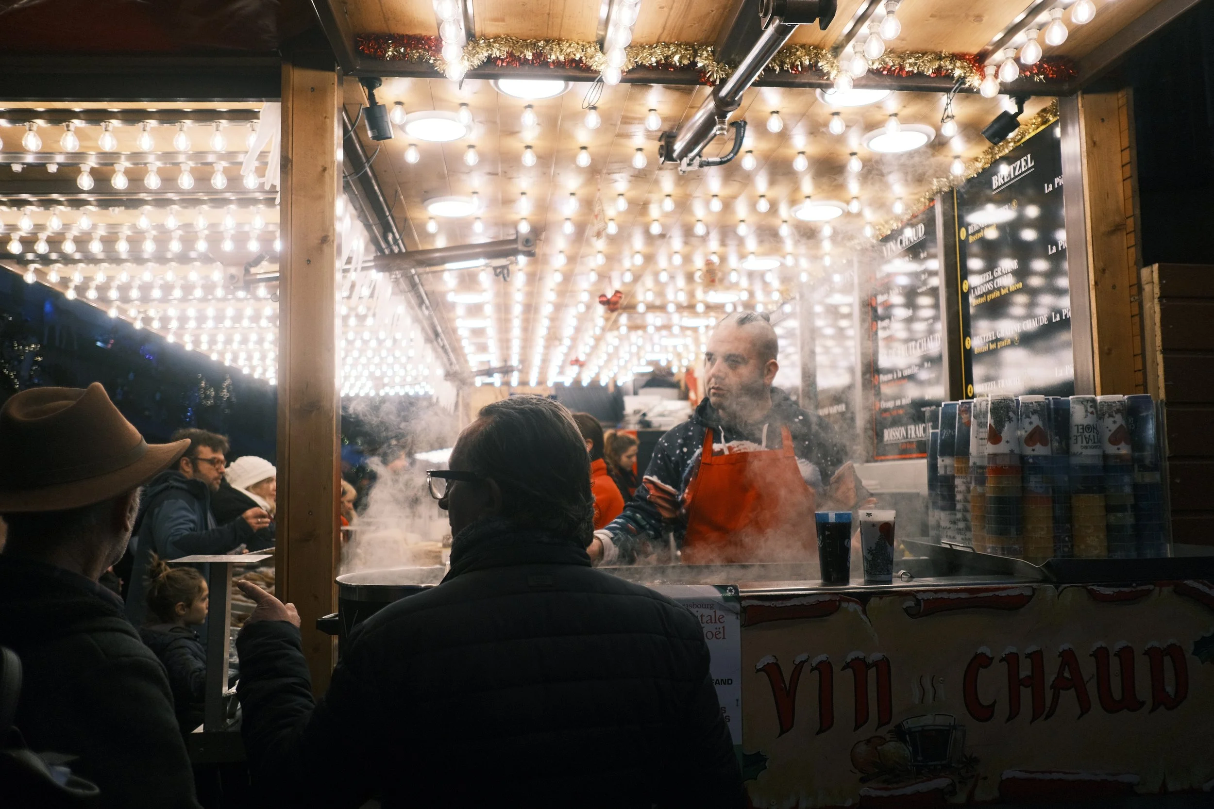 Stall de nourriture illuminé avec des lumières de Noël, où un vendeur prépare des plats fumants pour des clients lors d'un marché nocturne de Noël.