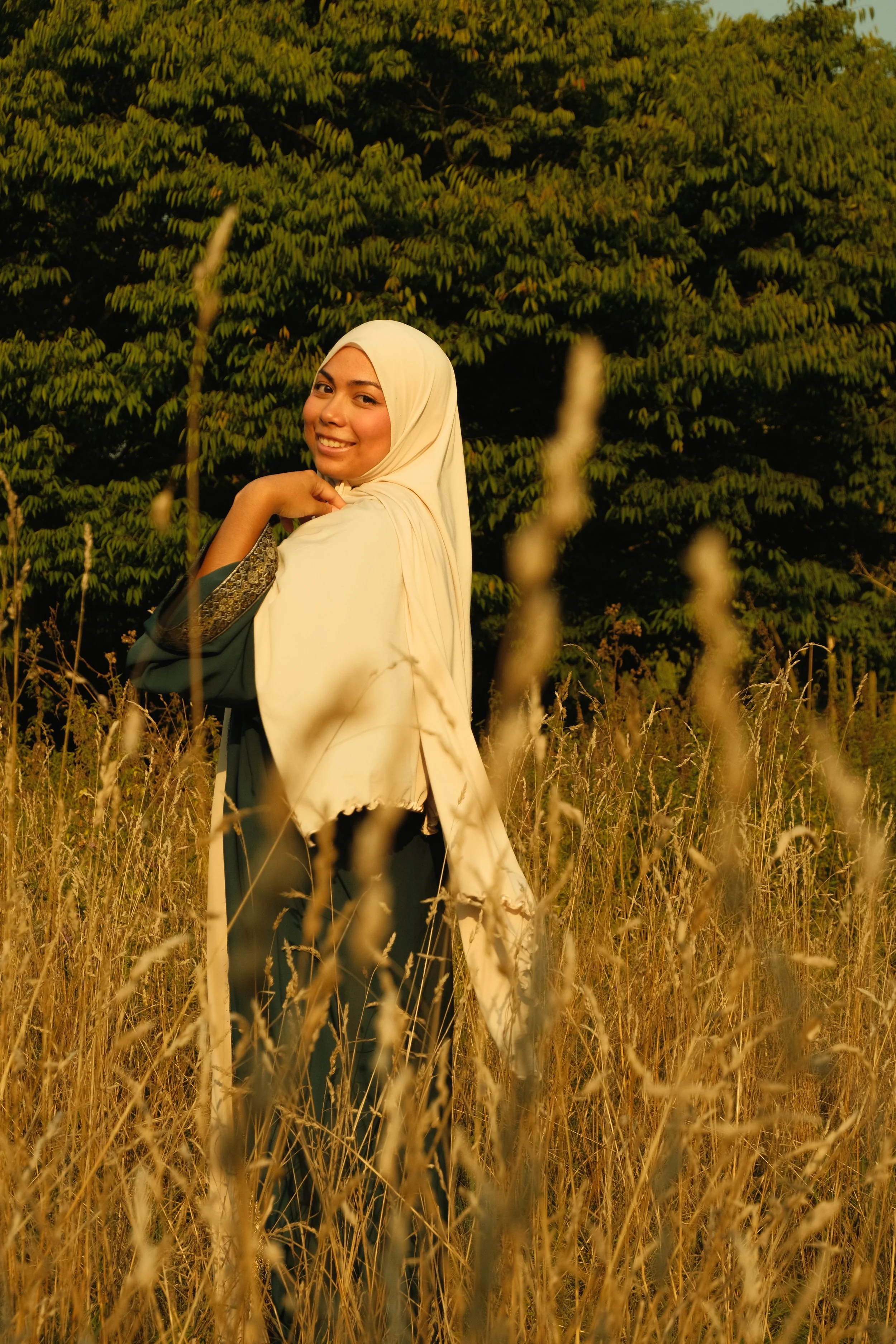 Femme souriante portant un hijab et une robe, debout dans un champ de hautes herbes, avec un arbre en arrière-plan.