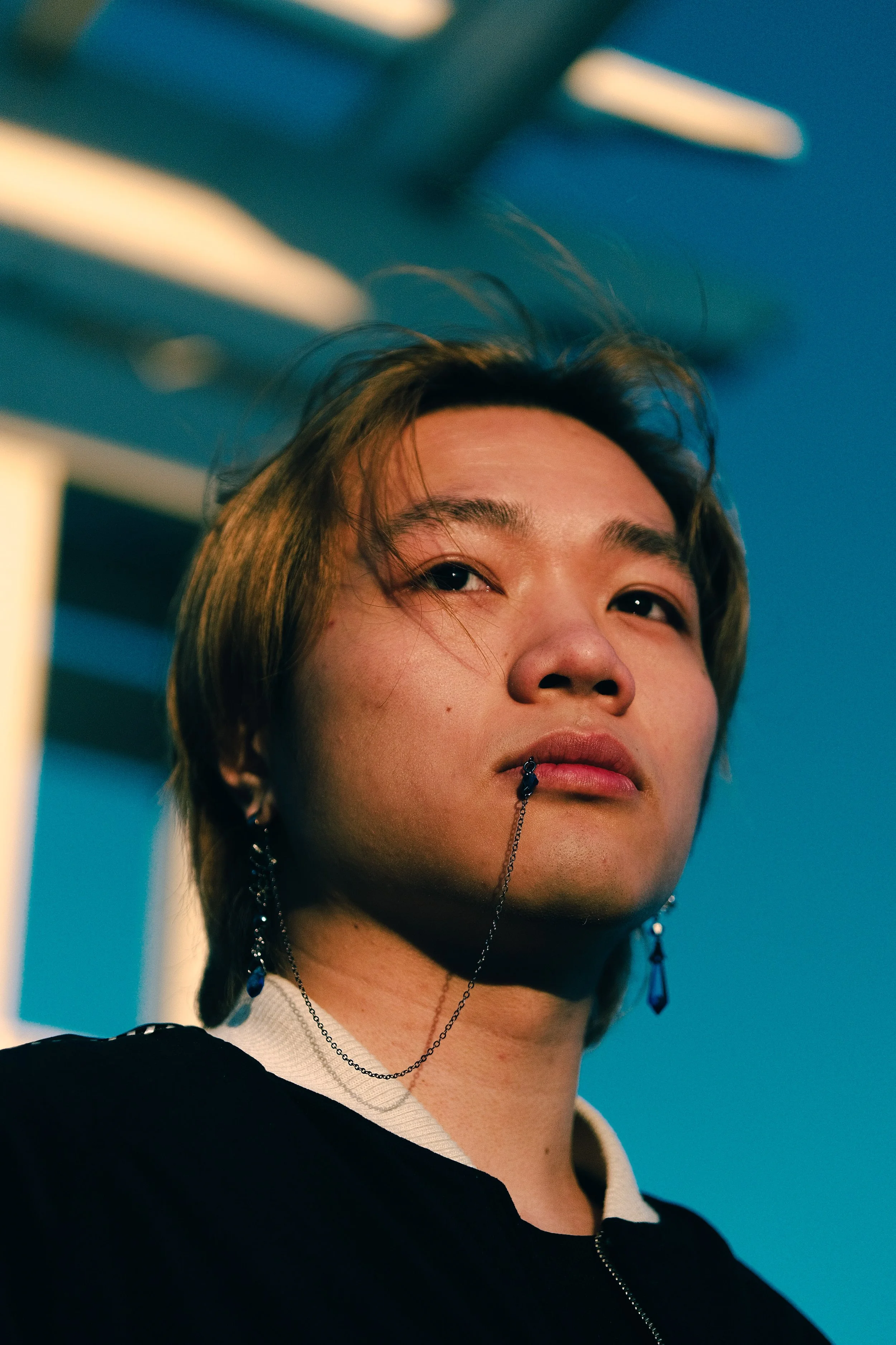 Portrait d'une personne avec des cheveux mi-longs, portant des boucles d'oreilles et un collier, optics lumineux, fond bleu.