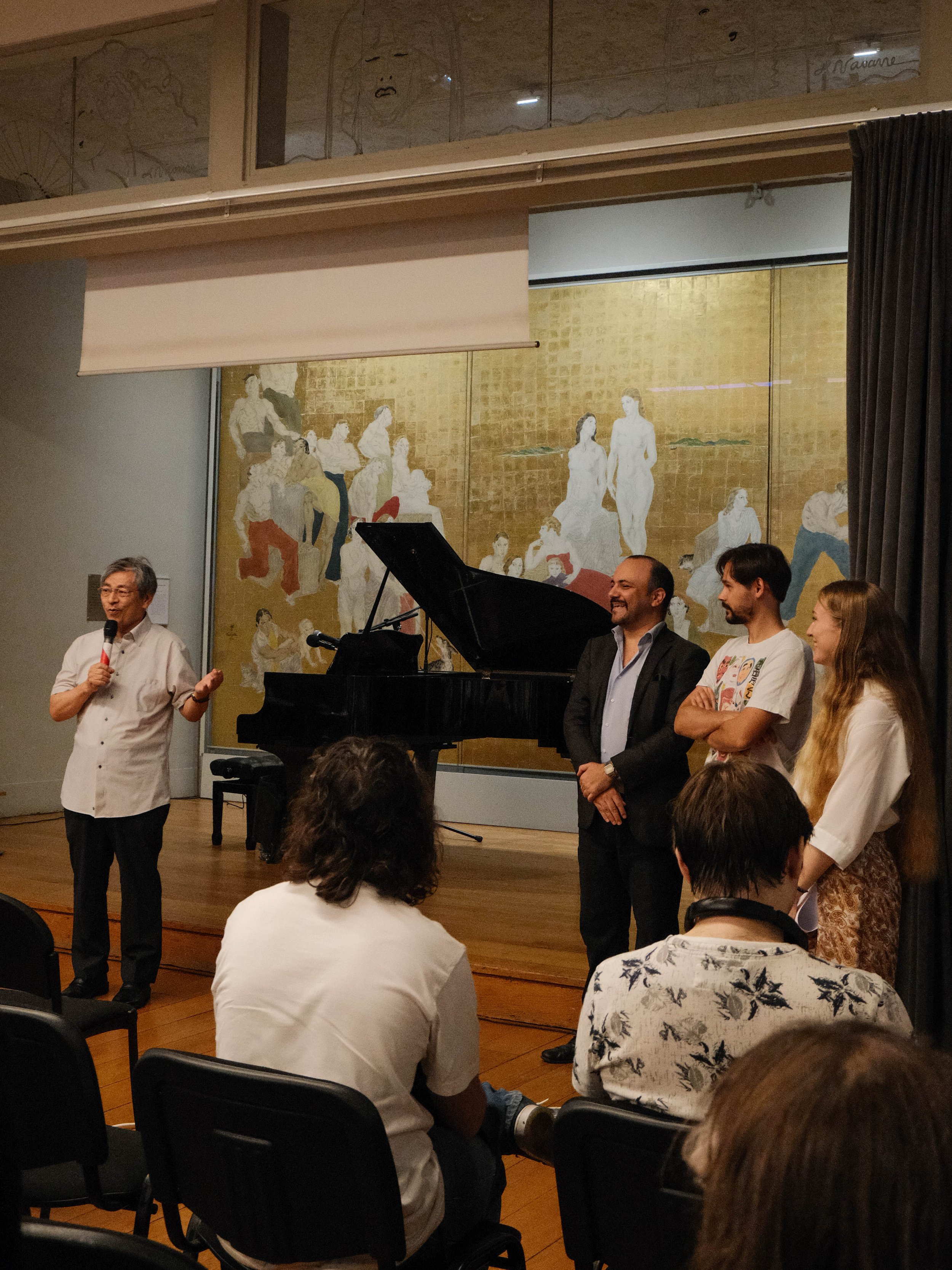 Une personne parle en arrière-plan avec un micro, devant un piano et un groupe de trois personnes qui écoutent. Il y a des spectateurs assis et un grand tableau avec une peinture en arrière-plan.