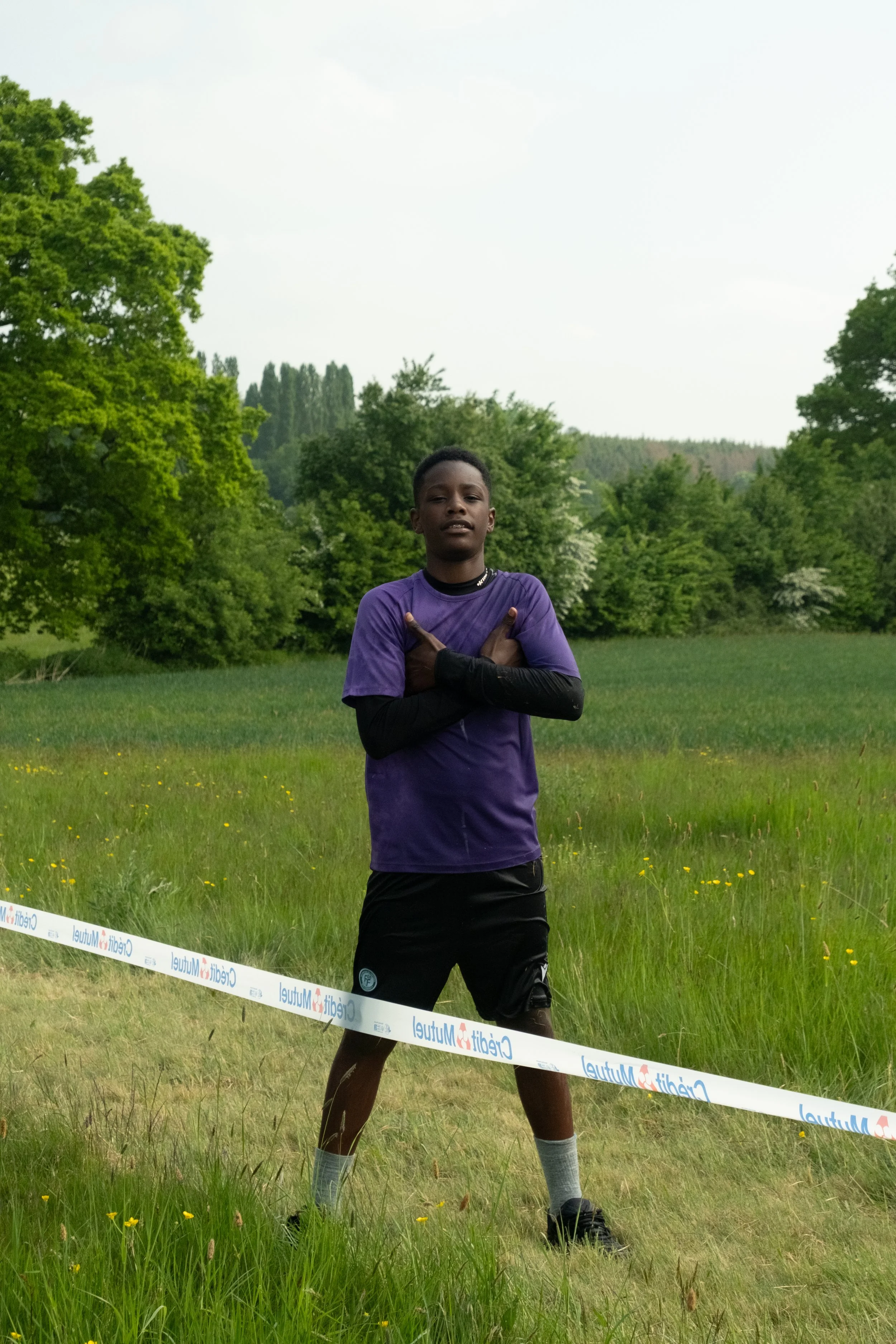 Un jeune homme debout dans une prairie verdoyante, portant un t-shirt violet, avec une chaîne autour du cou, et croisant ses bras. Il semble poser pour la photo dans un cadre naturel avec des arbres en arrière-plan.
