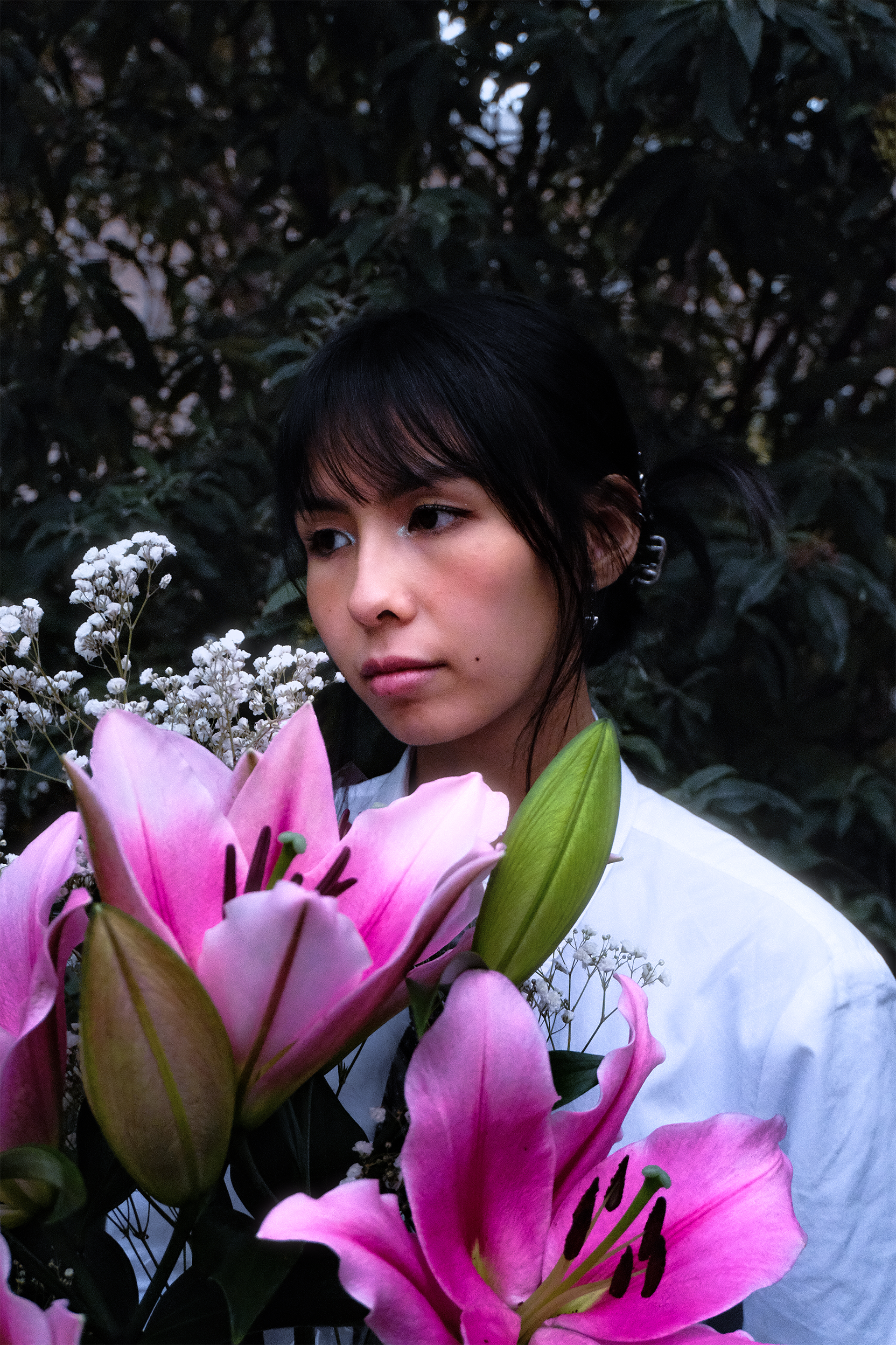 Une jeune femme avec des cheveux noirs, tenant un bouquet de fleurs, dont des lys roses et des petites fleurs blanches, dans un environnement naturel
