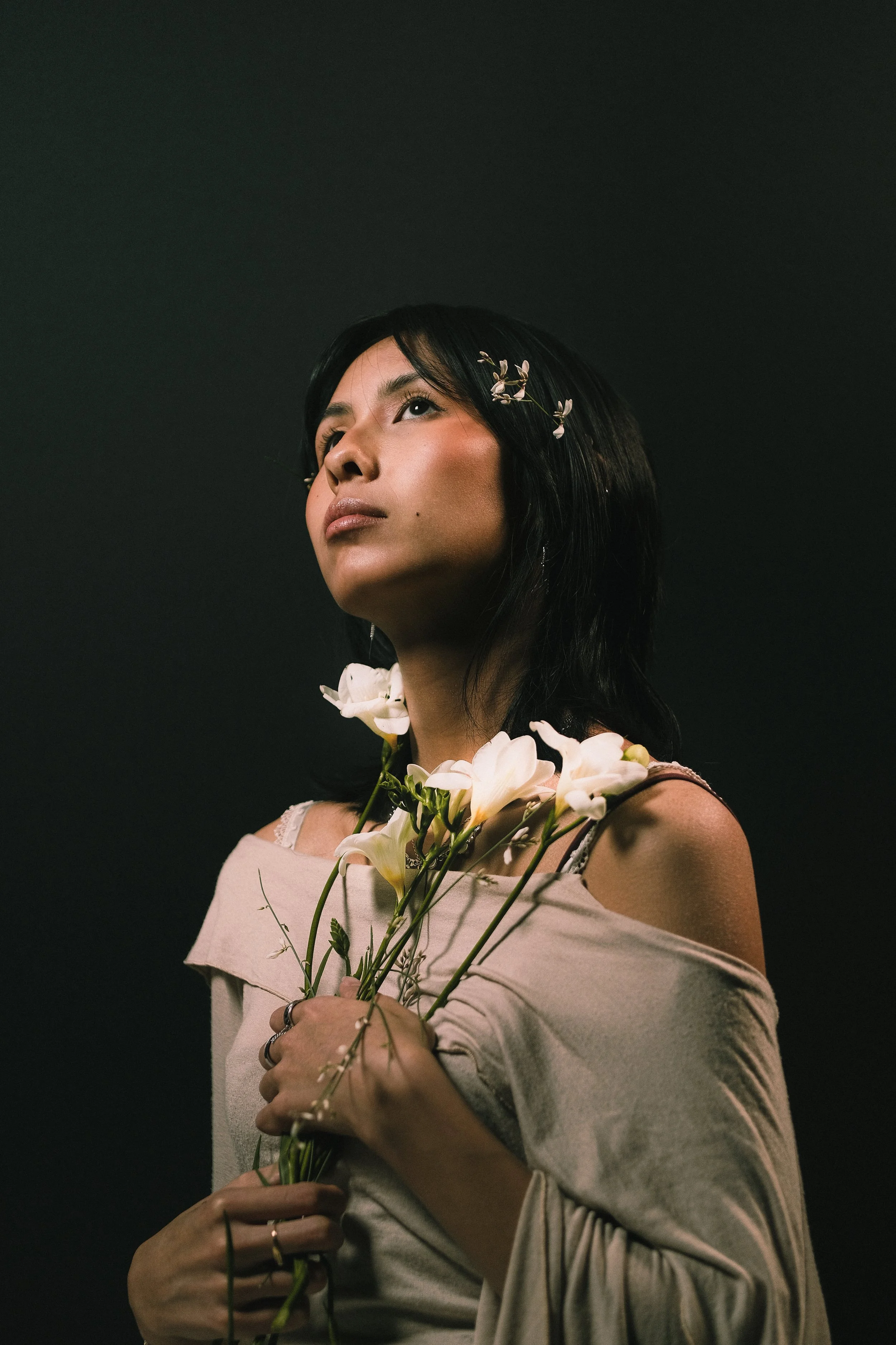 Une femme avec des fleurs blanches dans les cheveux, tenant un bouquet de fleurs blanches, regardant vers le haut, contre un fond sombre.