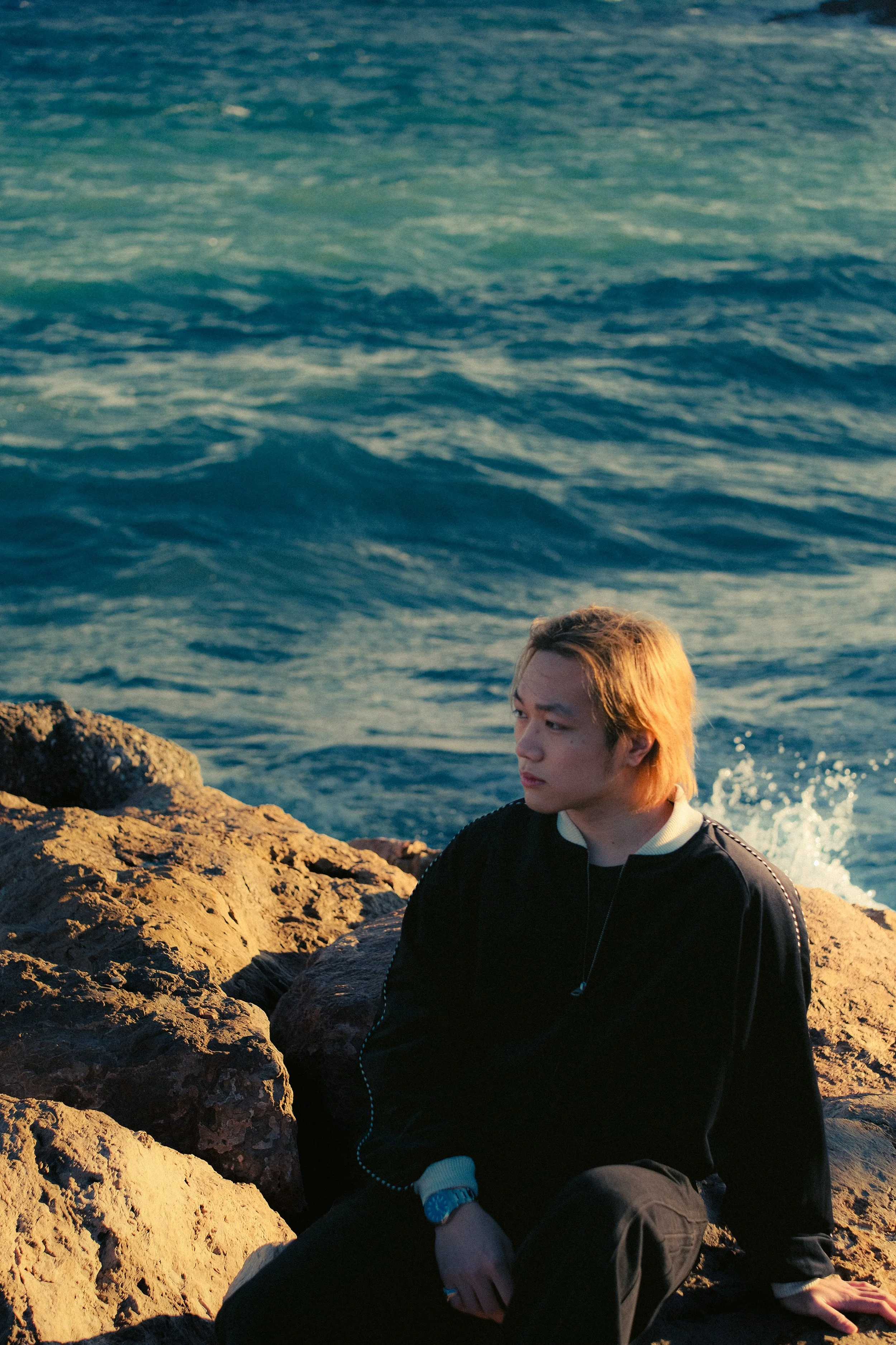 Jeune homme assis sur des rochers au bord de la mer, regardant pensivement l'horizon au coucher du soleil.