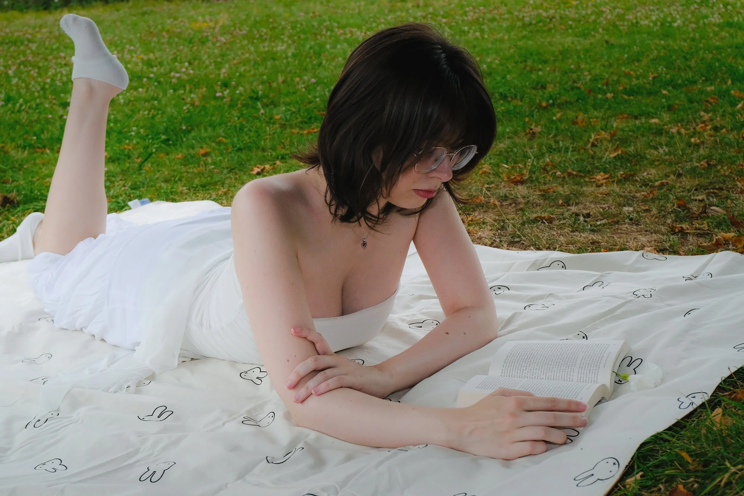 Une femme avec des lunettes, à moitié nue, allongée sur un drap blanc avec des dessins de lapins, lisant un livre en plein air sur de l'herbe dans un parc.