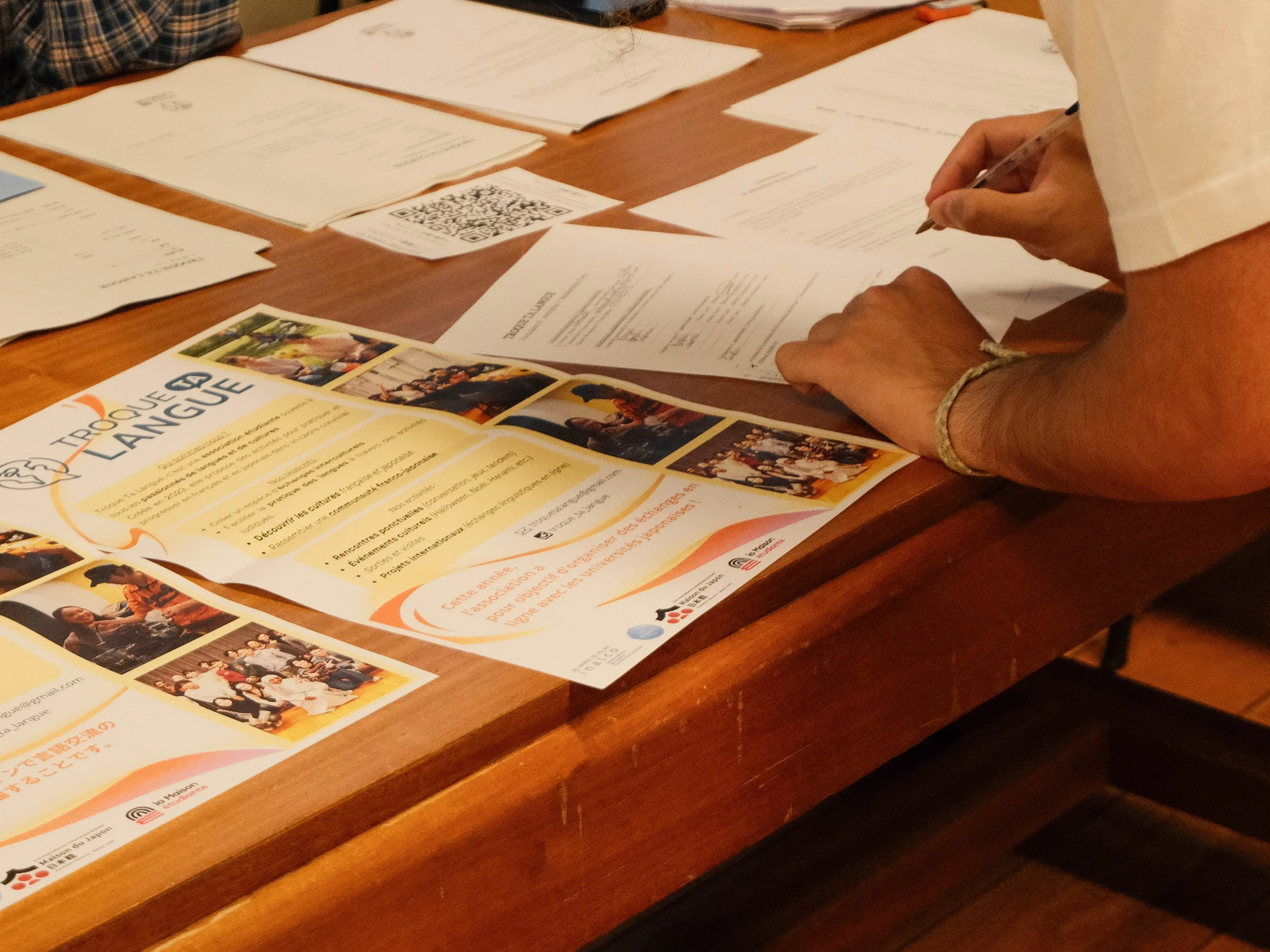 Une personne qui écrit sur une table en bois, entourée de différents papiers, brochures, et flyers, dont un sur l'apprentissage de la langue. La scène semble se dérouler lors d'une réunion ou d'une activité éducative.