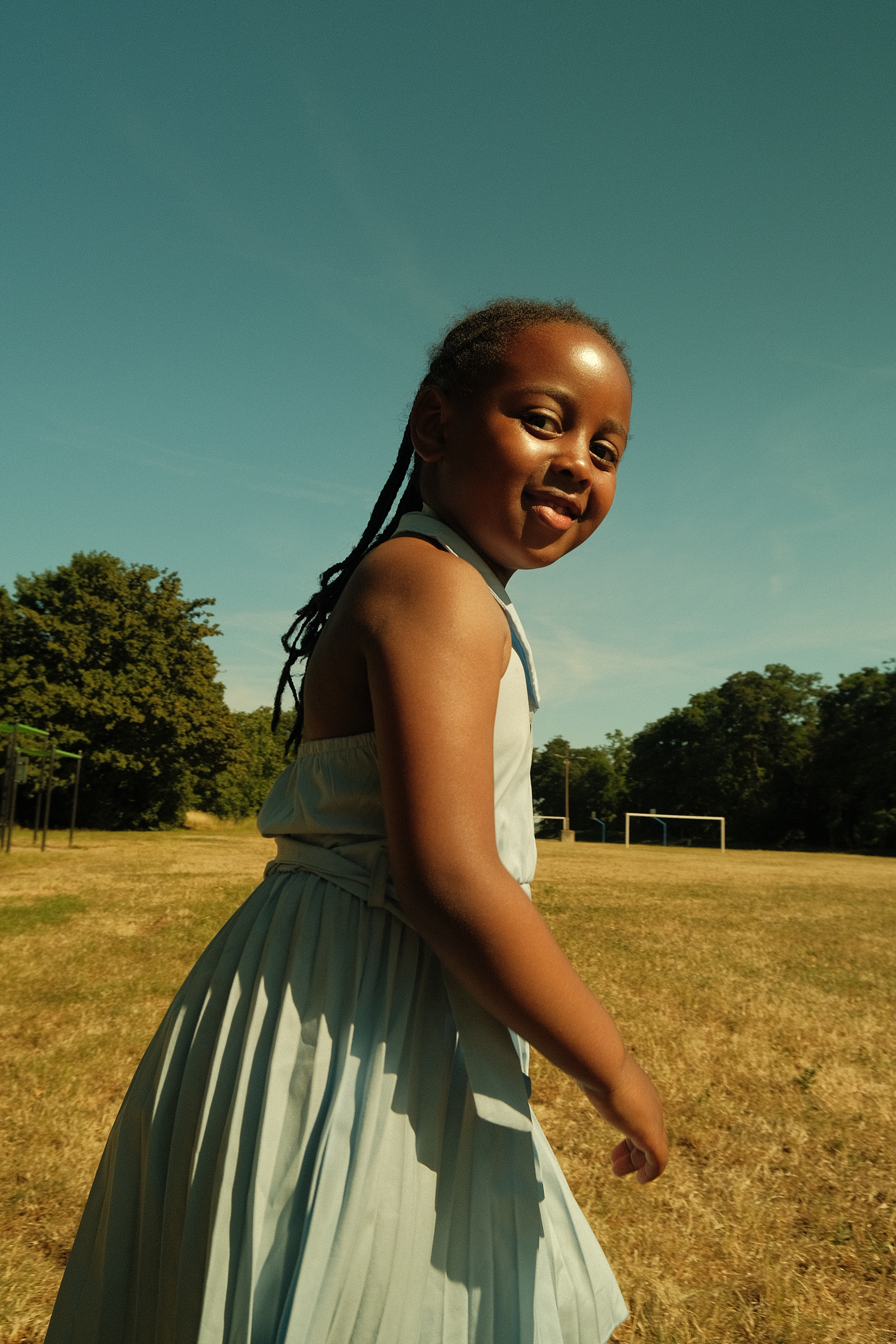 Une jeune fille souriante avec des dreadlocks, vêtue d'une robe à plis, marche dans un parc en plein jour. Le ciel est clair et il y a des arbres en arrière-plan.