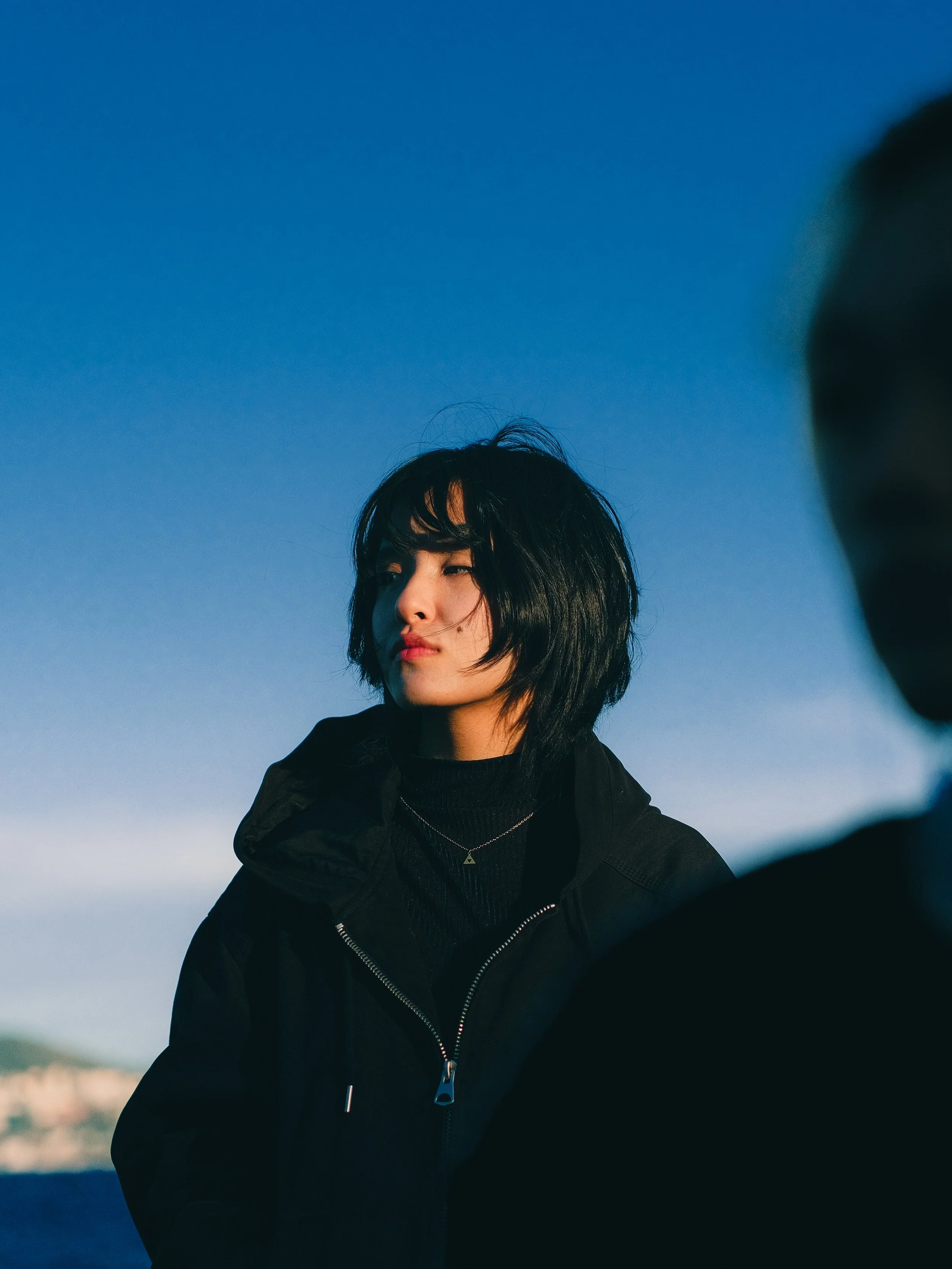 Jeune femme avec coupe de cheveux noire, portant une veste noire, regardant vers la gauche avec un fond de ciel bleu.