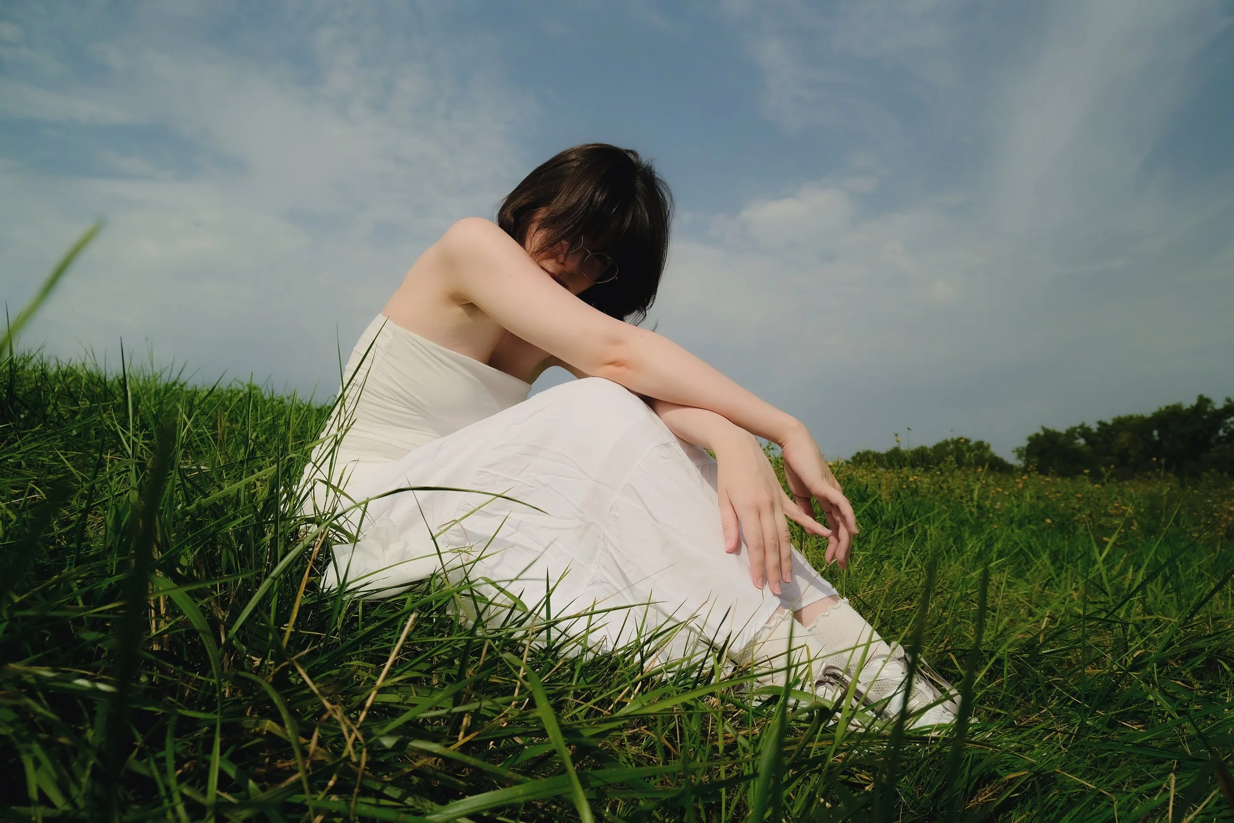 Femme assise dans l'herbe verte en plein air sous un ciel nuageux, portant une robe blanche, avec les cheveux noirs, regardant vers le sol, dans un paysage naturel avec des arbres au fond.