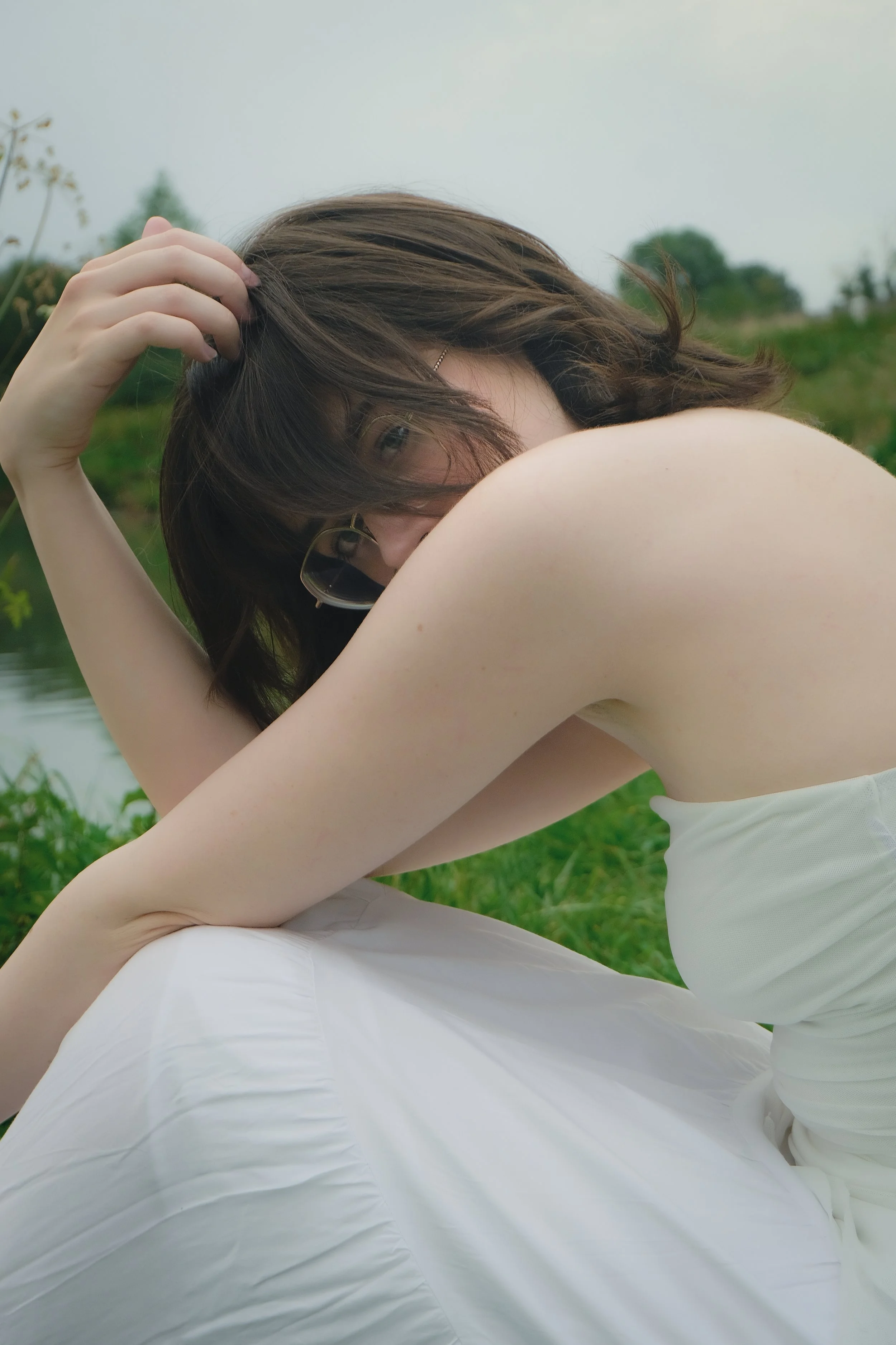 Jeune femme aux cheveux bruns, portant des lunettes, en robe blanche, assise près d'un étang, dans un environnement naturel.