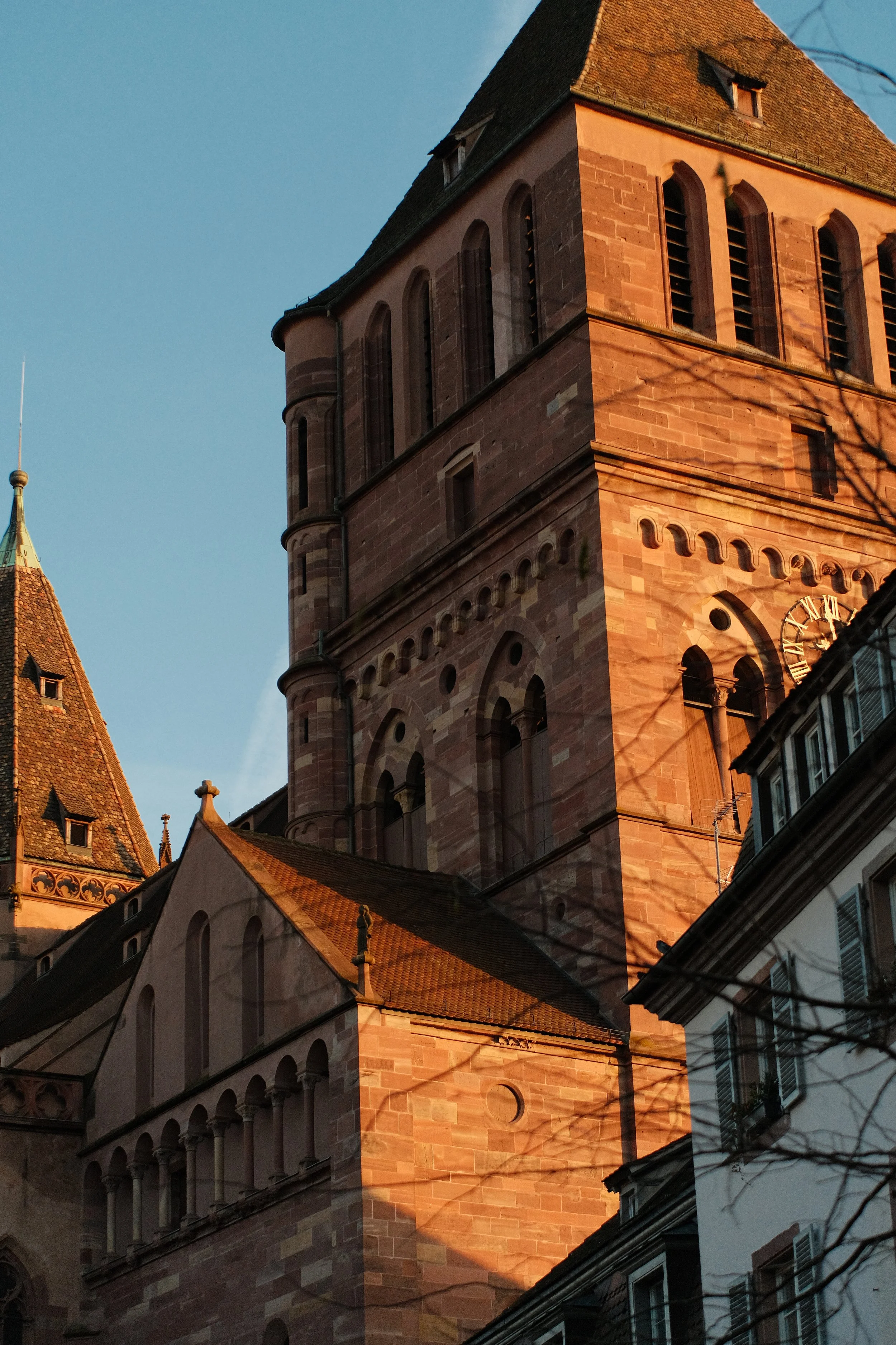 Une vieille église en pierre rose avec un clocher en forme de tour, sous un ciel bleu, éclairée par le soleil couchant.