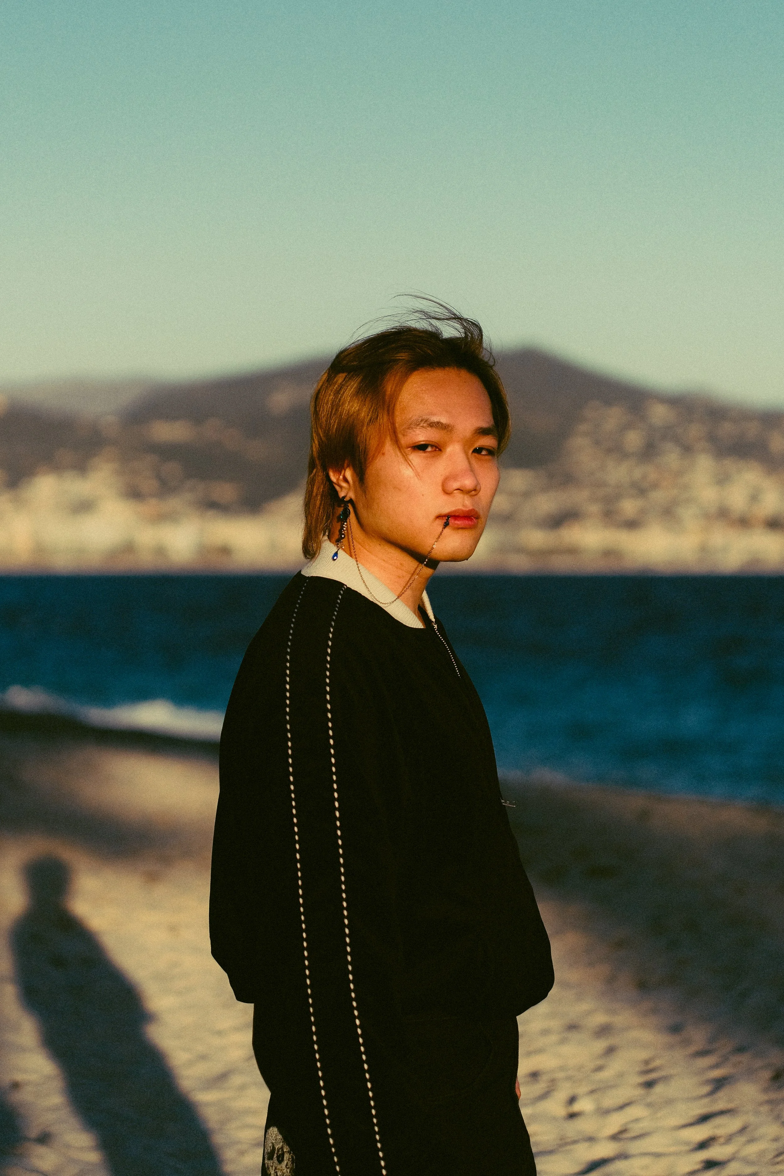 Jeune homme sur une plage au coucher du soleil, en vêtements noirs, avec des boucles d'oreilles, regardant vers la caméra avec un fond de mer et de montagnes.