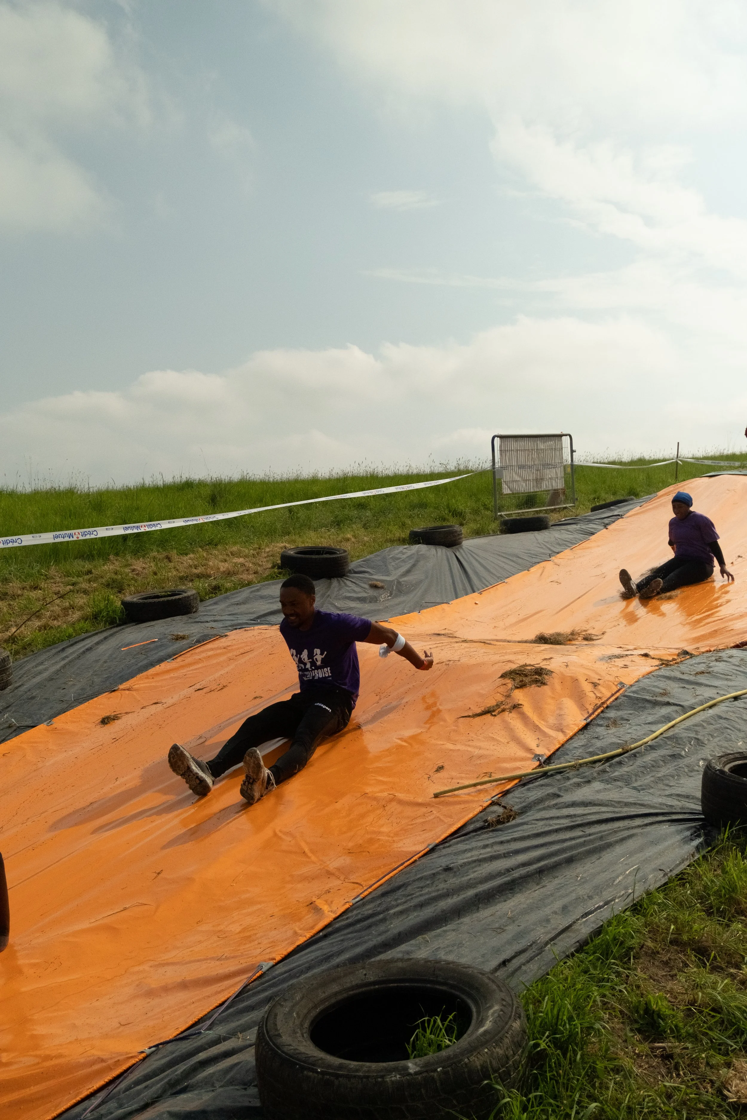 Deux enfants glissant sur une pente en plastique orange lors d'une activité en plein air, avec un ciel partiellement nuageux et un terrain herbeux en arrière-plan.