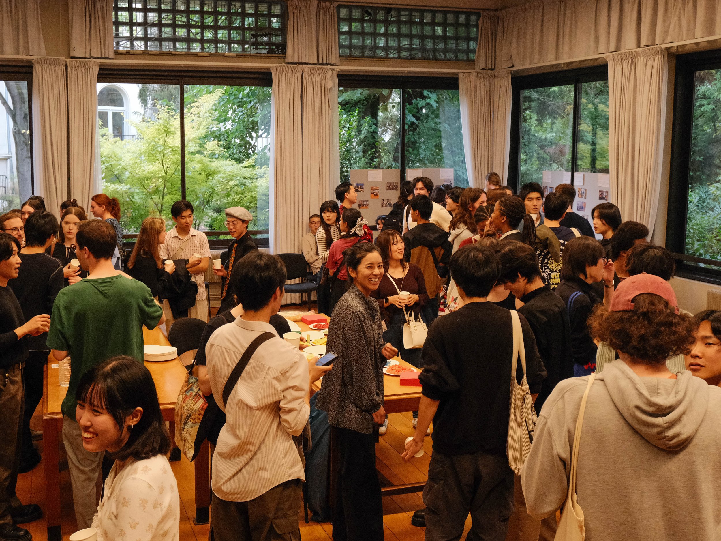 Une grande réunion ou un événement social avec beaucoup de personnes dans une salle lumineuse avec de grandes fenêtres et des rideaux blancs. Des gens discutent, prennent des selfies et se tiennent autour d'une table avec des petits plats.