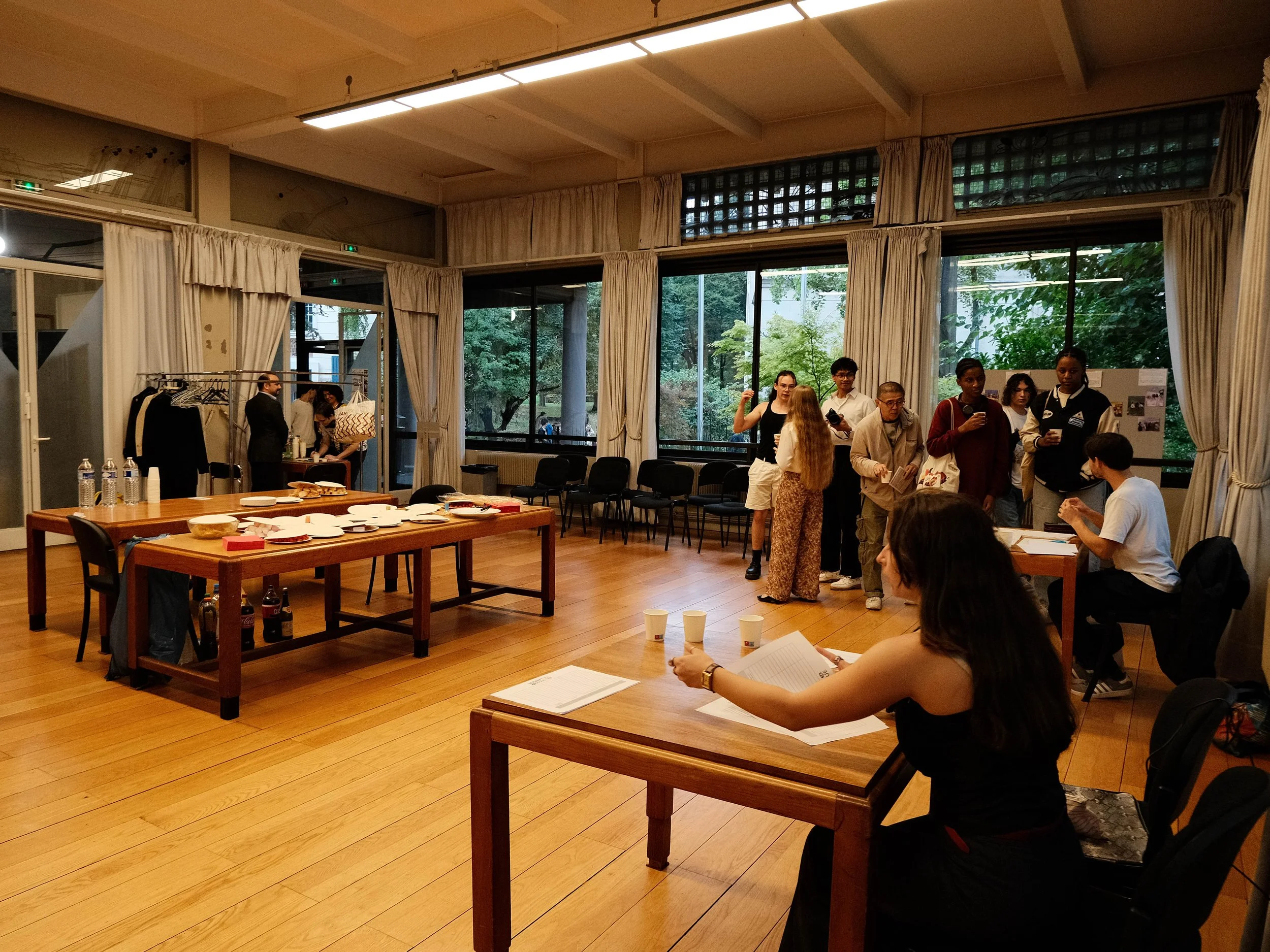 Une salle de réunion avec plusieurs personnes en train de discuter, une table avec des bouteilles d'eau et des assiettes vides, et un espace avec des vêtements suspendus. Les fenêtres offrent une vue du jardin extérieur.