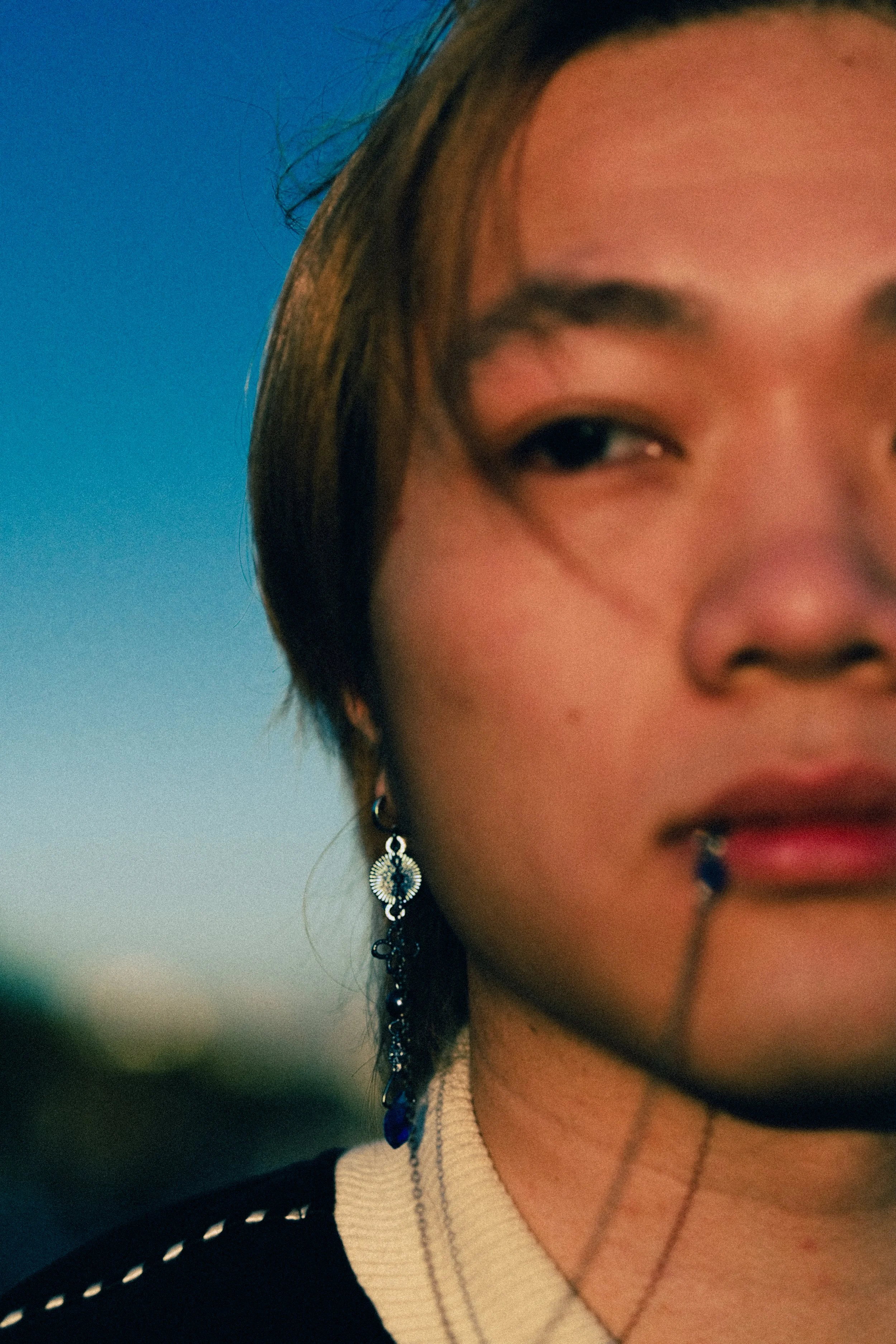 Un gros plan du visage d'une femme, avec un fond de ciel bleu. Elle porte une boucle d'oreille ornementale et a une lèvre perforée avec un piercing. La photo est prise lors du coucher du soleil ou au crépuscule.