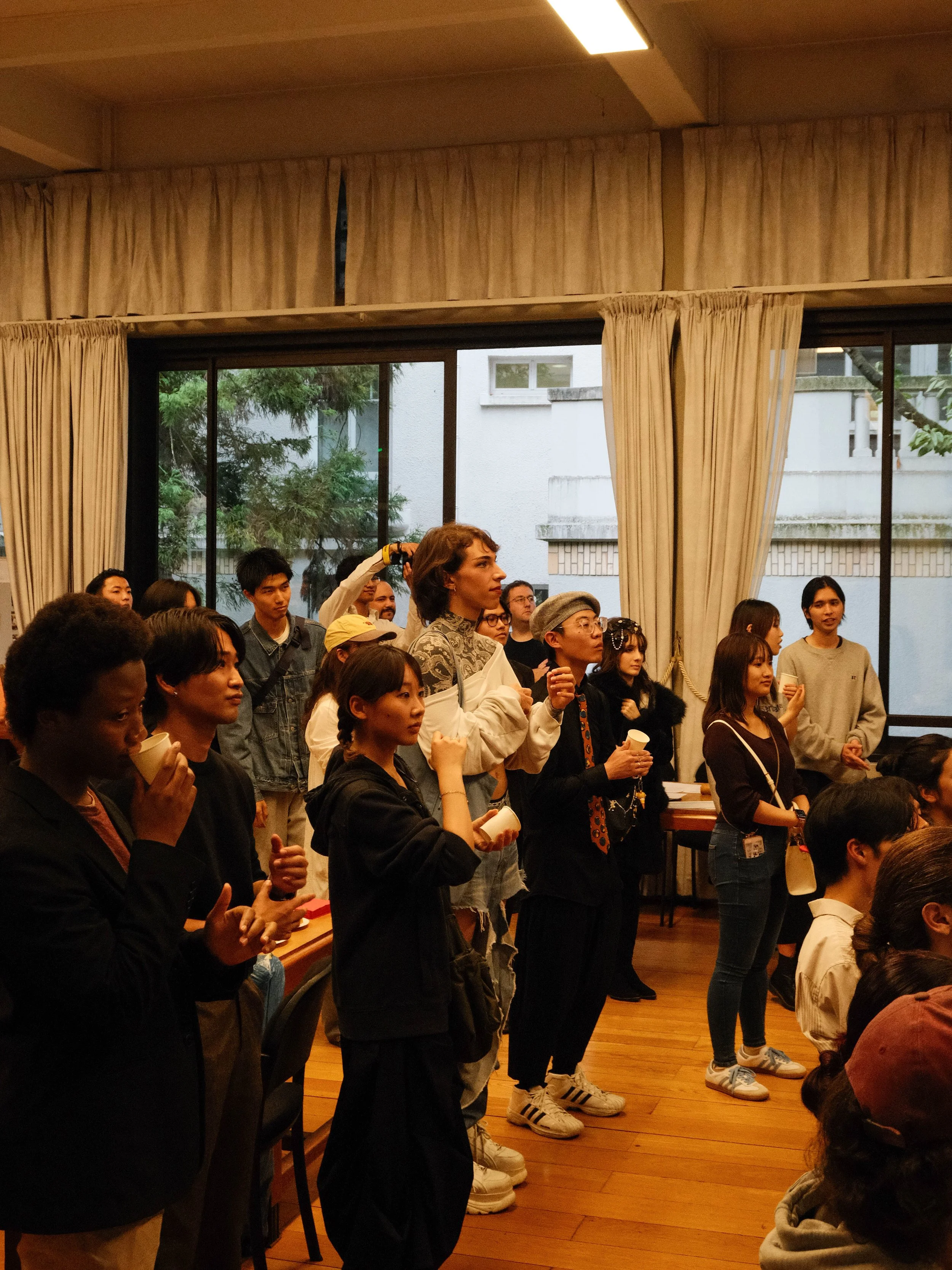 Groupe de personnes debout dans une salle, certaines tenant des tasses en papier, regardant vers l'avant, avec un décor intérieur comprenant des fenêtres avec des rideaux.