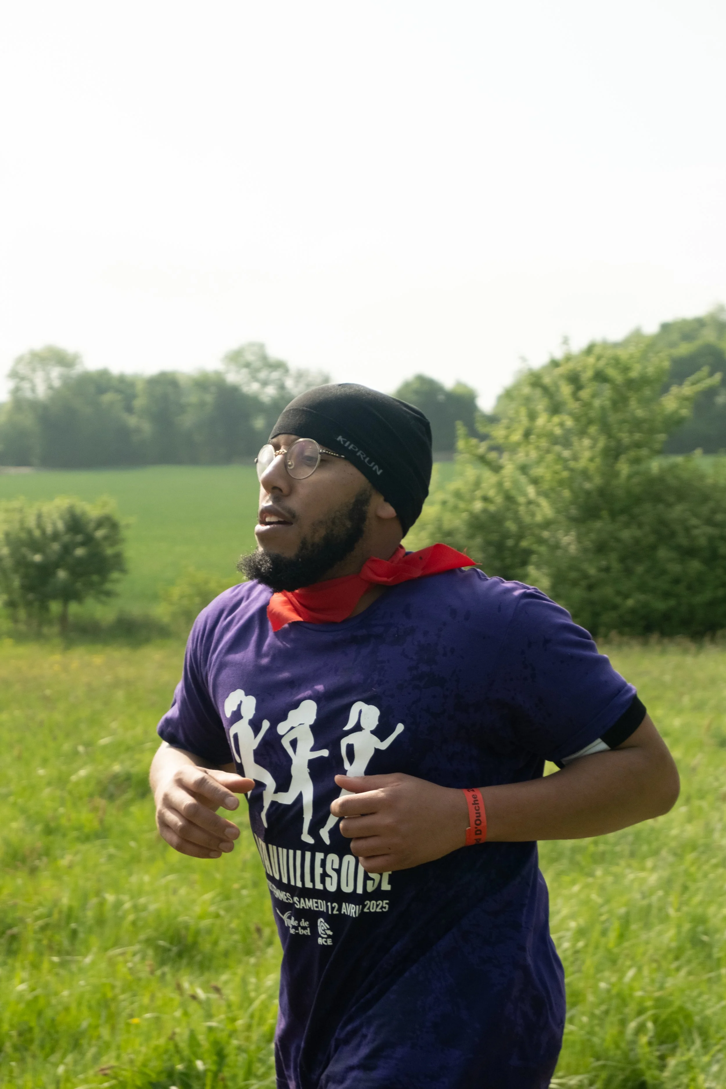 Un homme court dans un champ vert avec des arbres en arrière-plan, portant un t-shirt violet avec un graphique de marcheurs, un bandeau noir, des lunettes, un bracelet orange et un foulard rouge autour du cou.