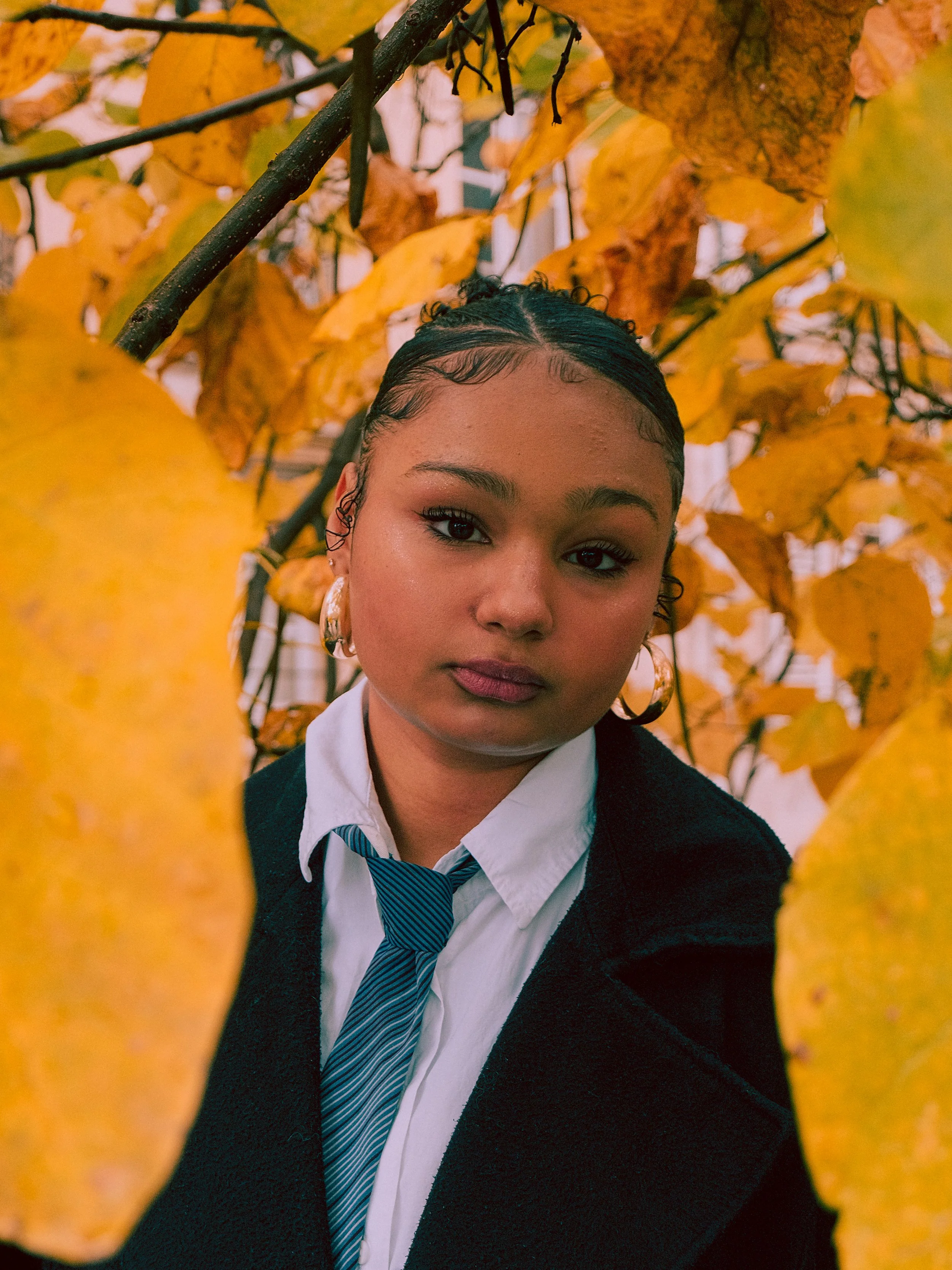 Jeune femme avec des cheveux bouclés courts, portant une chemise blanche, une cravate bleue, et un blazer noir, entourée de feuilles jaunes d'automne.