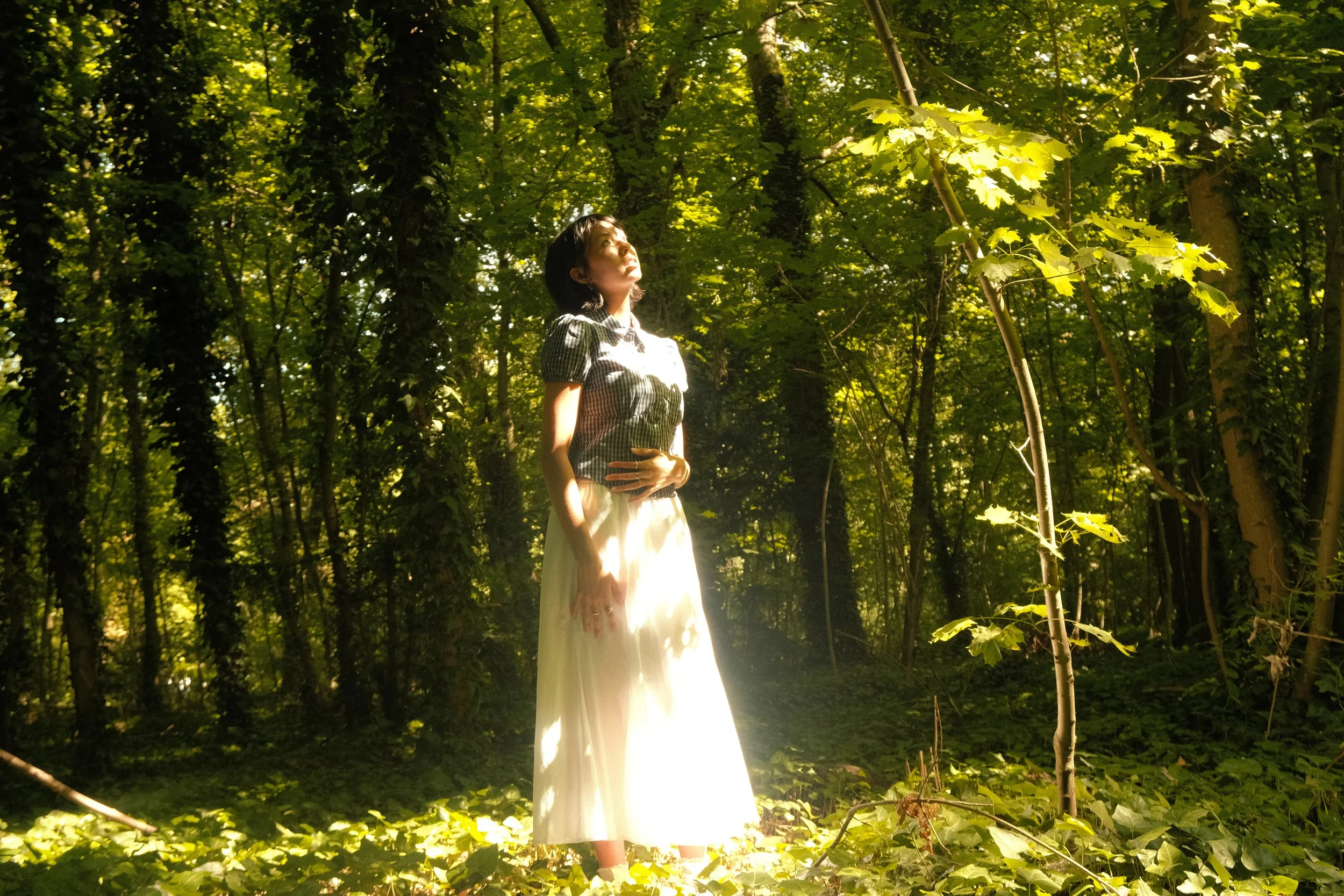 Une femme debout dans une forêt verdoyante, éclairée par la lumière du soleil filtrant à travers les arbres.