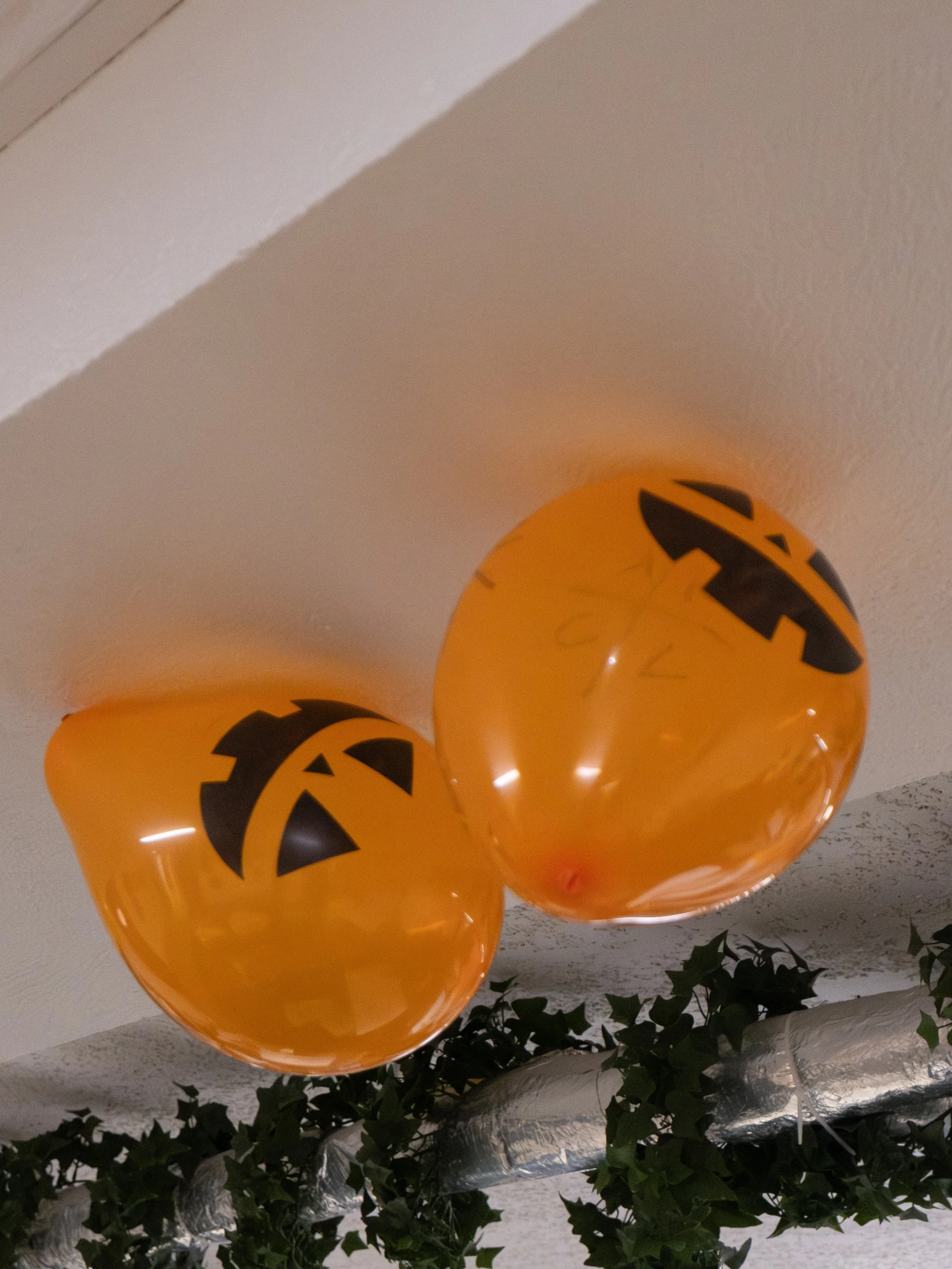 Deux ballons orange avec des visages de citrouille d'Halloween suspendus au plafond, entourés de plantes vertes.