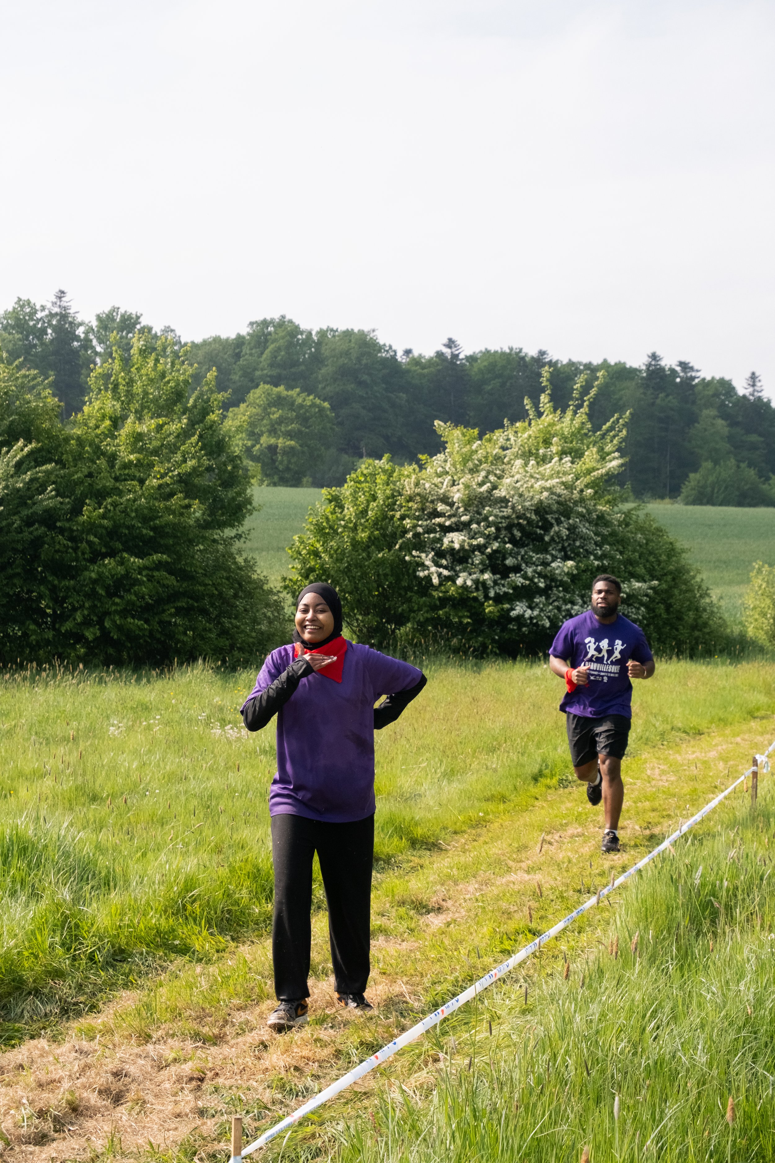 Deux personnes courent dans un champ vert, un fond de collines, deux buissons floraux, une femme souriante en avant-plan et un homme derrière elle, tous deux en t-shirt violet.